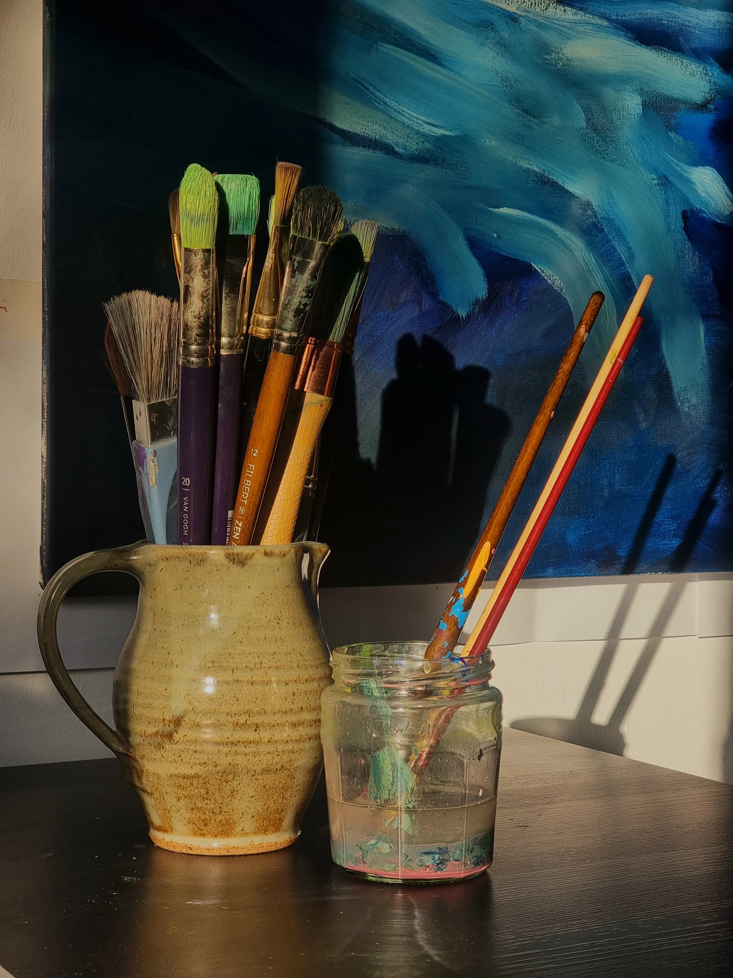 Paintbrushes in a ceramic pitcher and a jar of water on a table, with an abstract blue painting in the background.