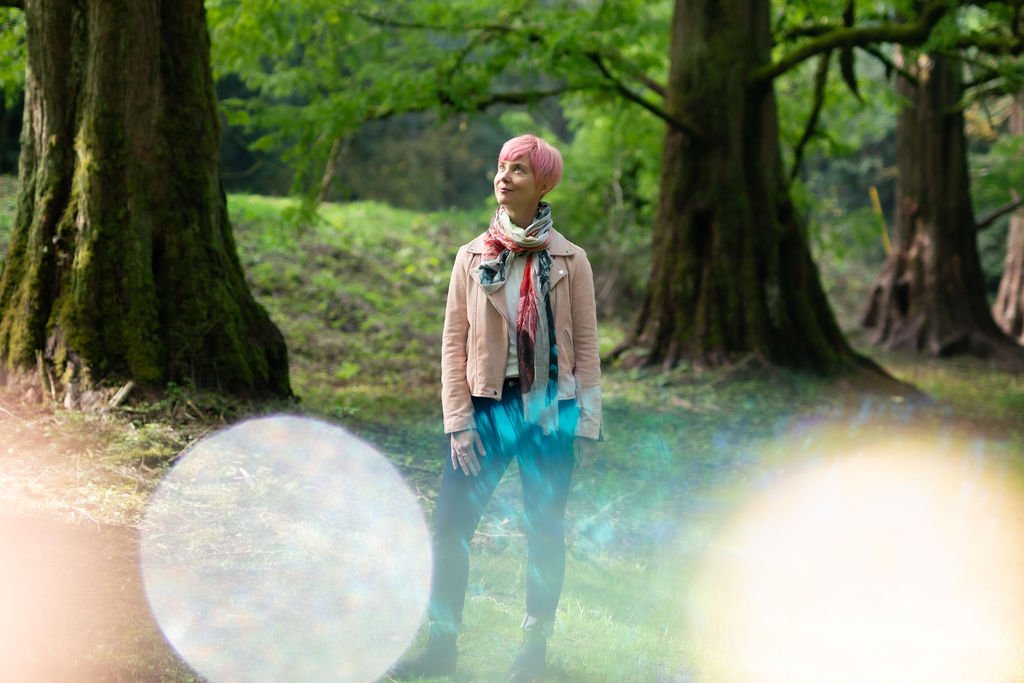 A woman with pink hair standing in a lush green forest, looking up, wearing a beige jacket, colorful scarf, and jeans, surrounded by trees.