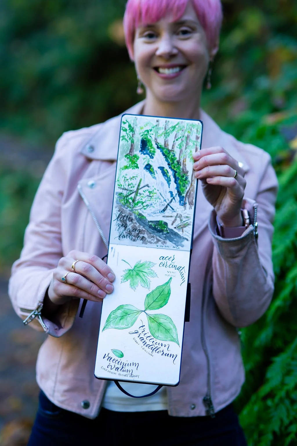 A woman smiles while holding her watercolor sketchbook filled with botanical illustrations.