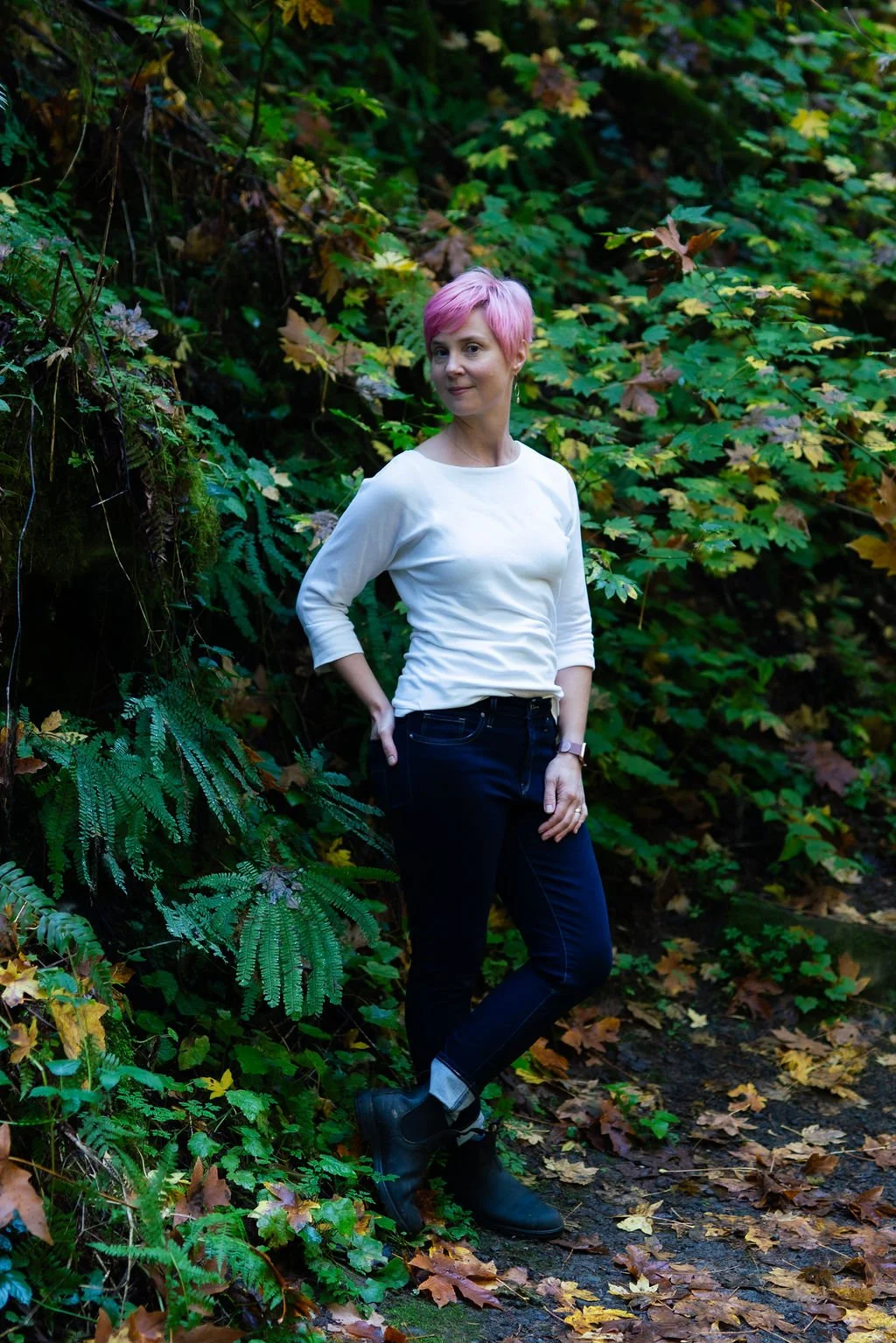 A woman with pink hair standing in a forest surrounded by green and yellow leaves, wearing a white long-sleeve shirt, black jeans, and black boots.