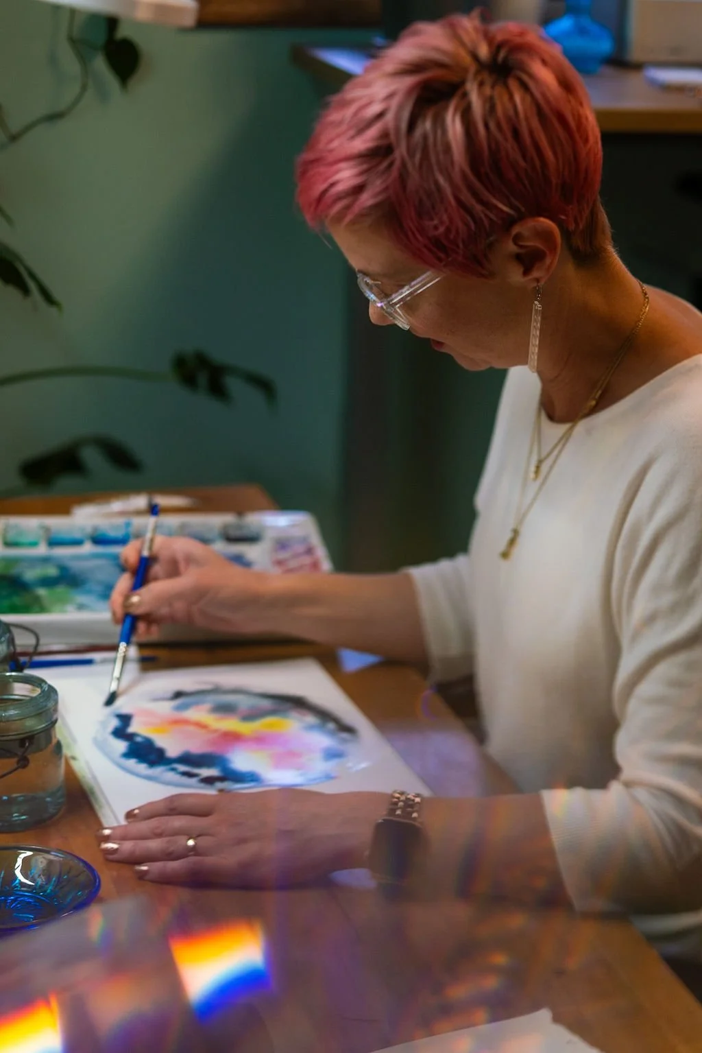 A woman sits at her painting table with a paintbrush and watercolor.