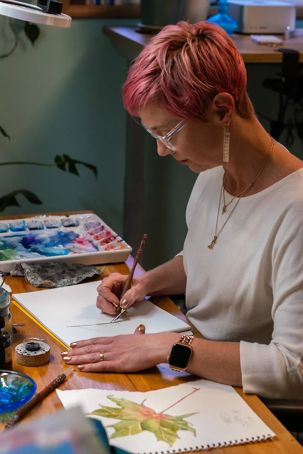 Woman with pink hair and glasses painting with a fine brush at a wooden desk, watercolor paints and drawings of a dragonfly on the paper.