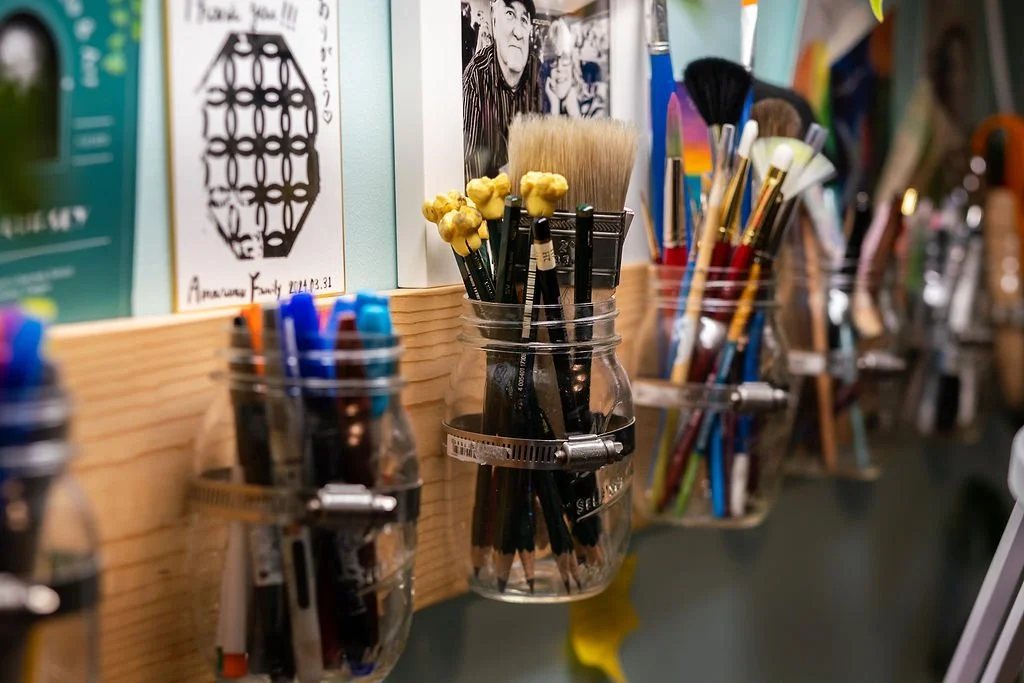 A collection of art brushes and pens stored in glass jars attached to a wooden board, with artwork and photos on the wall behind.
