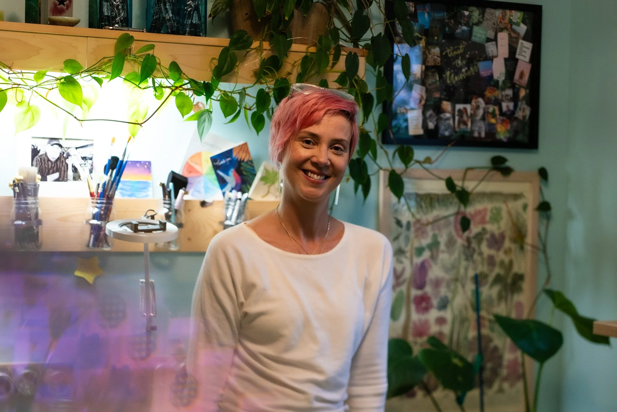 An artist woman stands in her illustration studio, surrounded by art supplies and plants. She has pink hair and a white sweater, with sparkle glasses on her head.