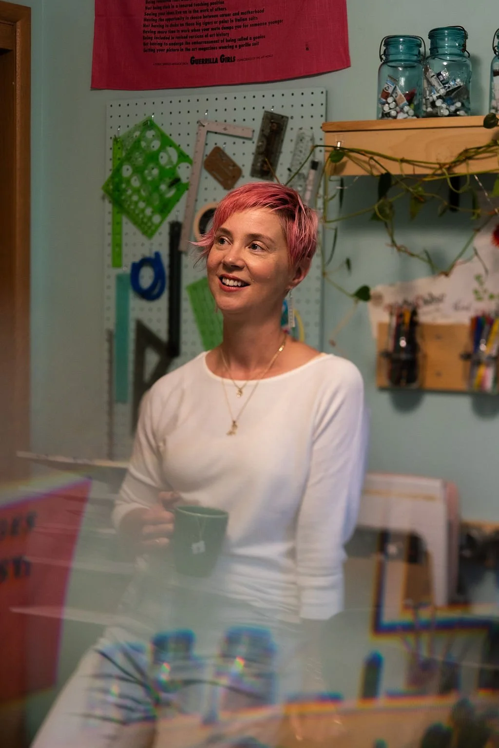Molly Chidsey, a woman with pink pixie haircut, standing in her art studio holding a mug of tea.