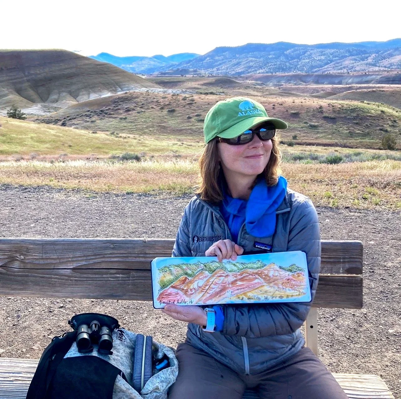 A woman in a green baseball hat holds her sketchbook of a watercolor illustration in the Oregon Outback painted hills park.
