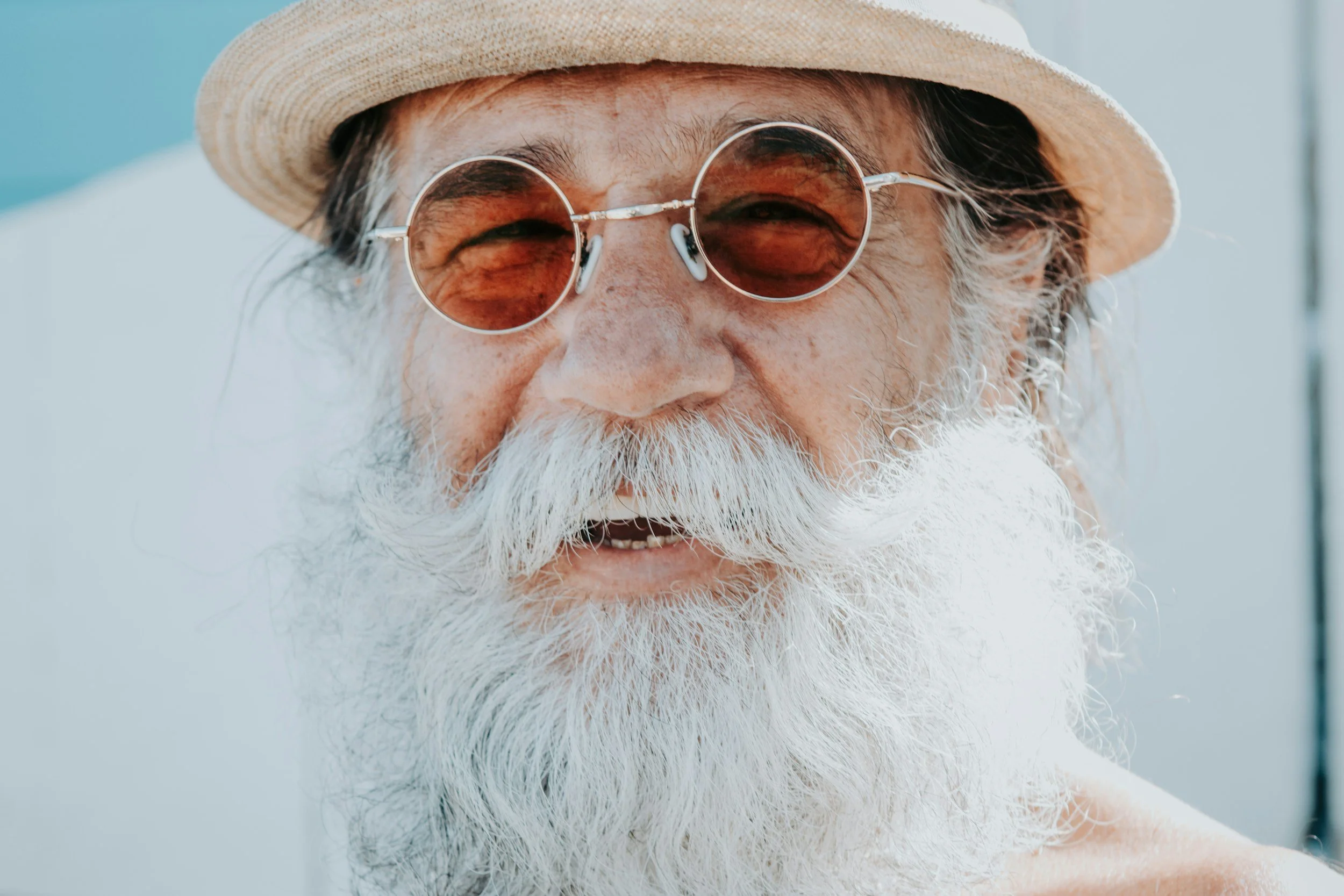 Close-up of an elderly man with a white beard and moustache, wearing round sunglasses and a straw hat, smiling outdoors.