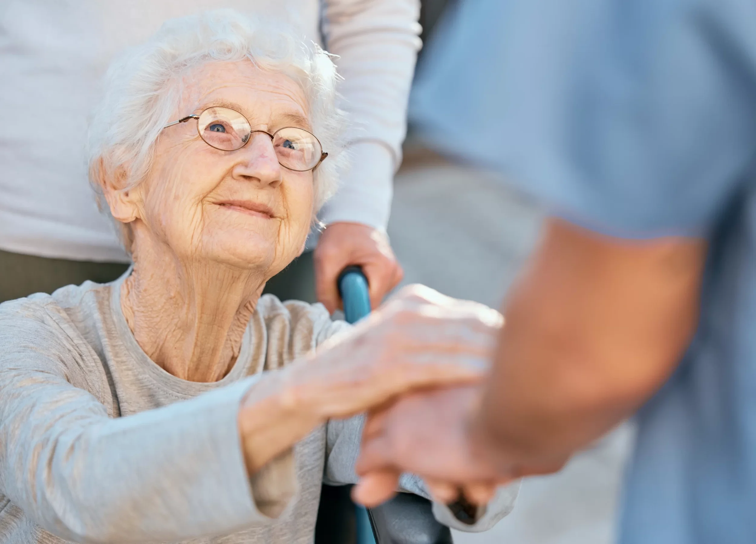 An elderly woman with glasses smiling while holding a caregiver's hand, with a walker nearby.