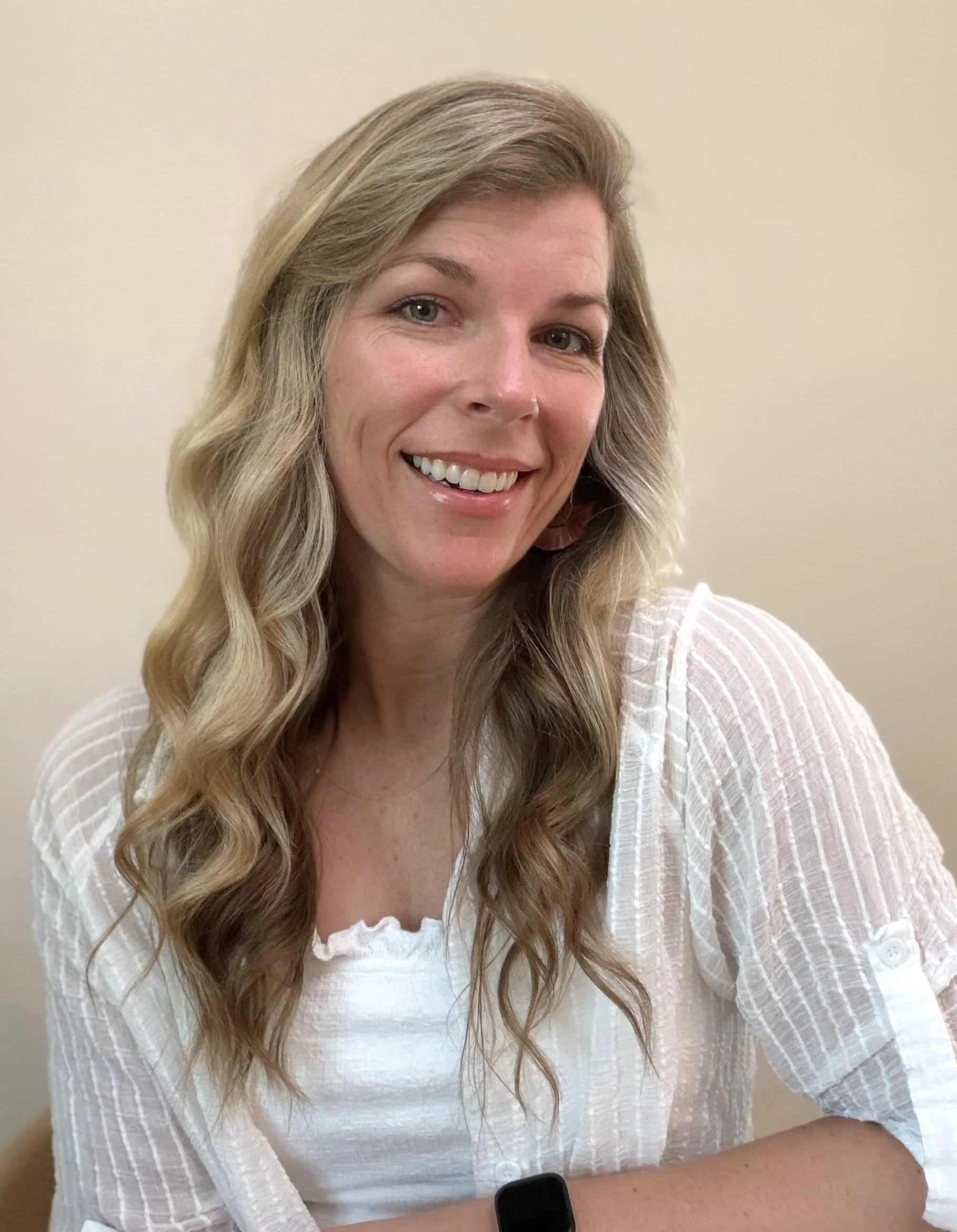 A woman with long wavy blonde hair, wearing a white top and striped cover-up, smiling with her mouth open, against a plain light-colored wall.