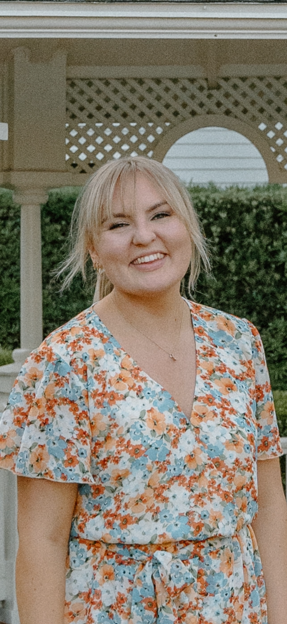 A woman with blonde hair wearing a floral dress, smiling while standing outdoors near a lattice fence and greenery.