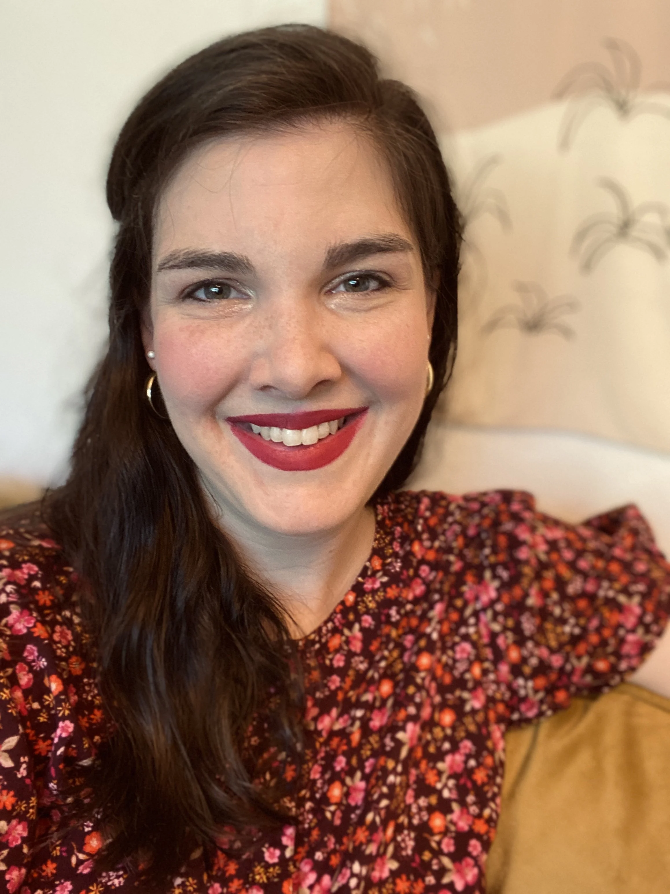 Close-up of a woman's face smiling, with long dark hair, red lipstick, and wearing earrings, in a floral-patterned top, sitting on a yellow couch with a decorative wall featuring birds in the background.