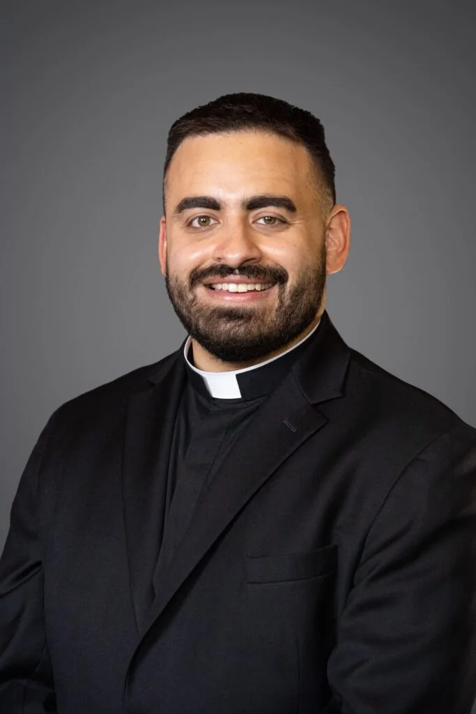 Silhouettes of a group of people standing on a beach during sunset or dusk, some holding phones and taking pictures.