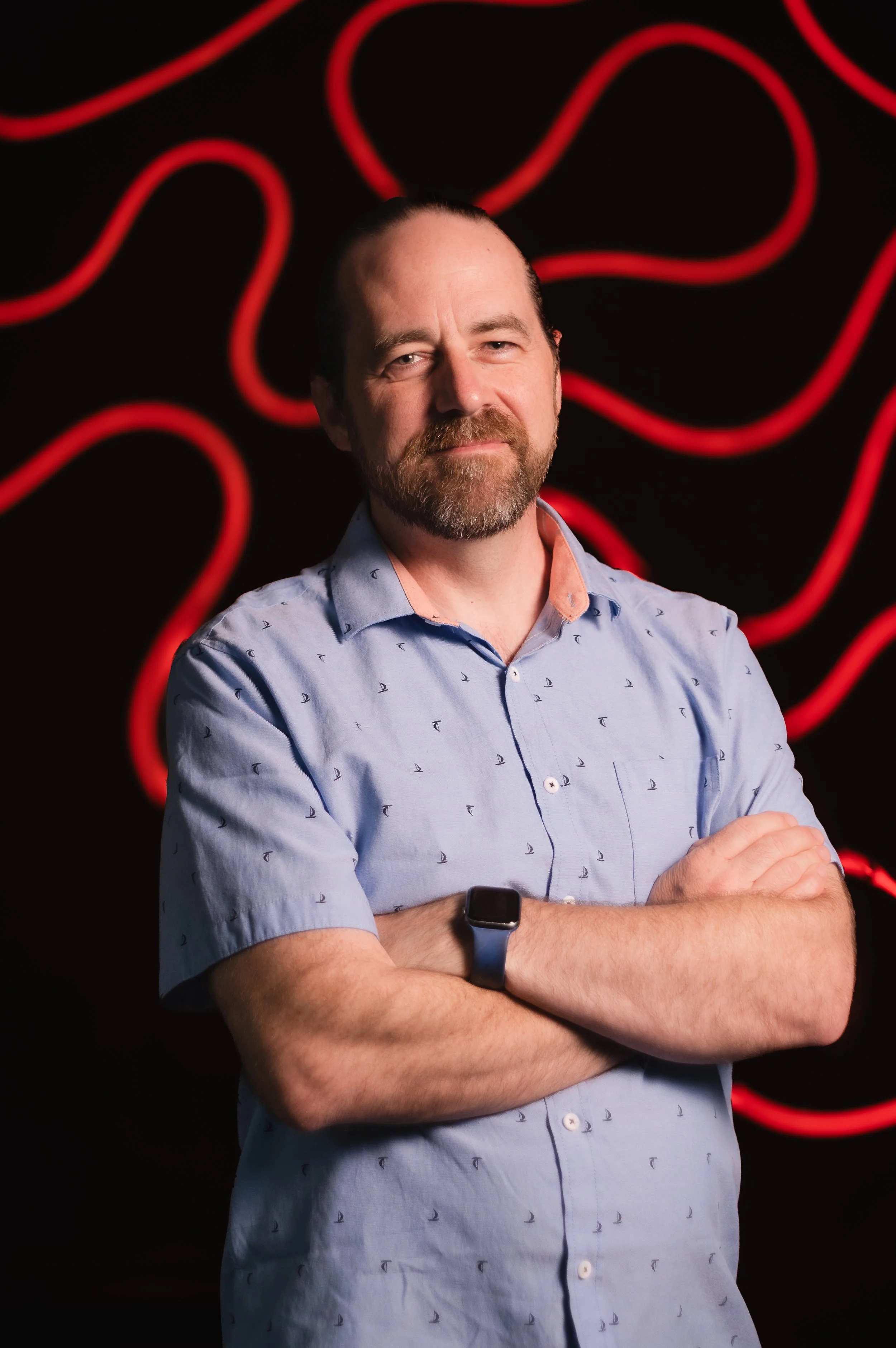 Man in a gray checked shirt with crossed arms standing in front of a colorful mosaic wall.