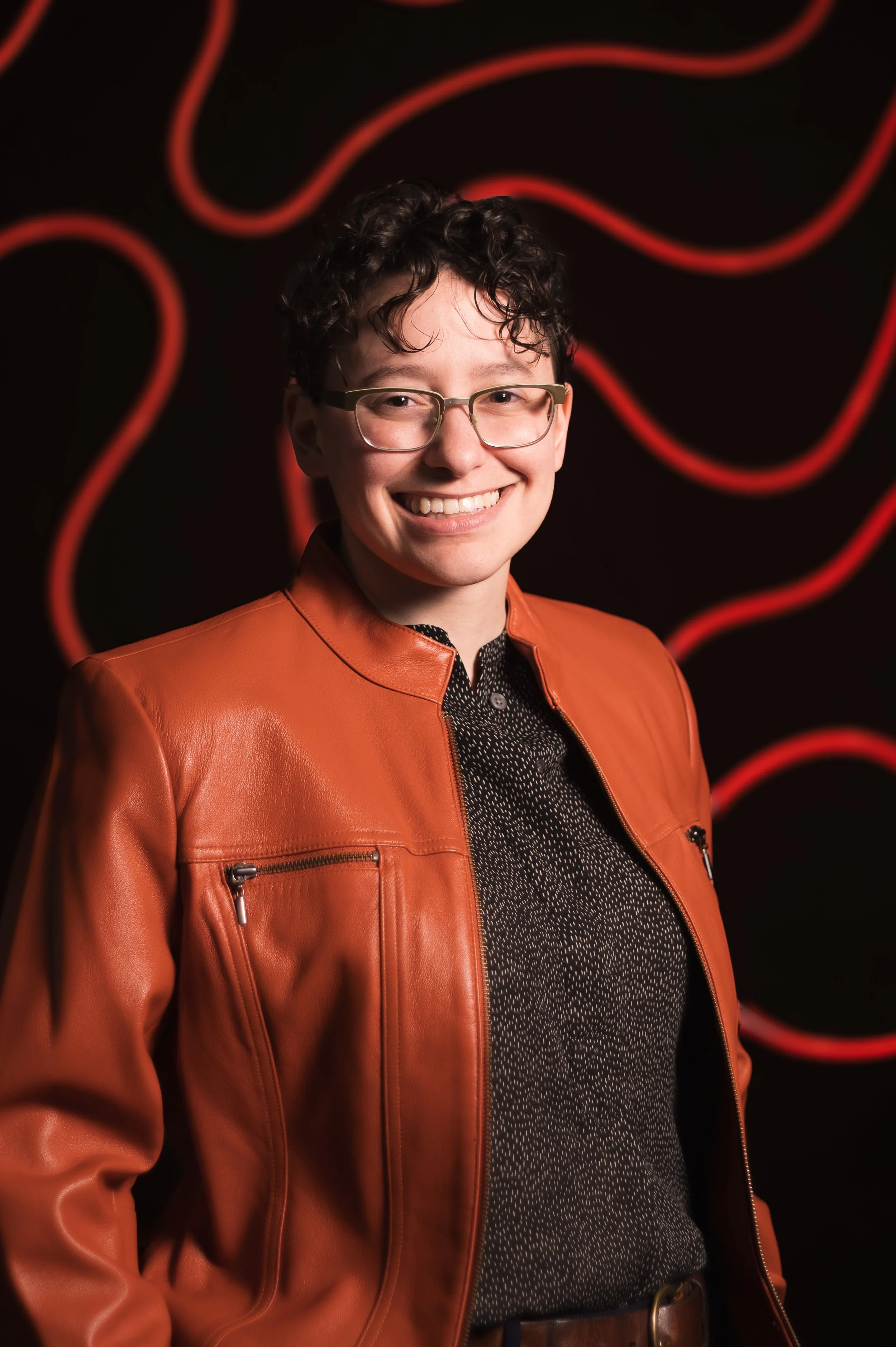Smiling person with glasses in front of a colorful tiled wall.