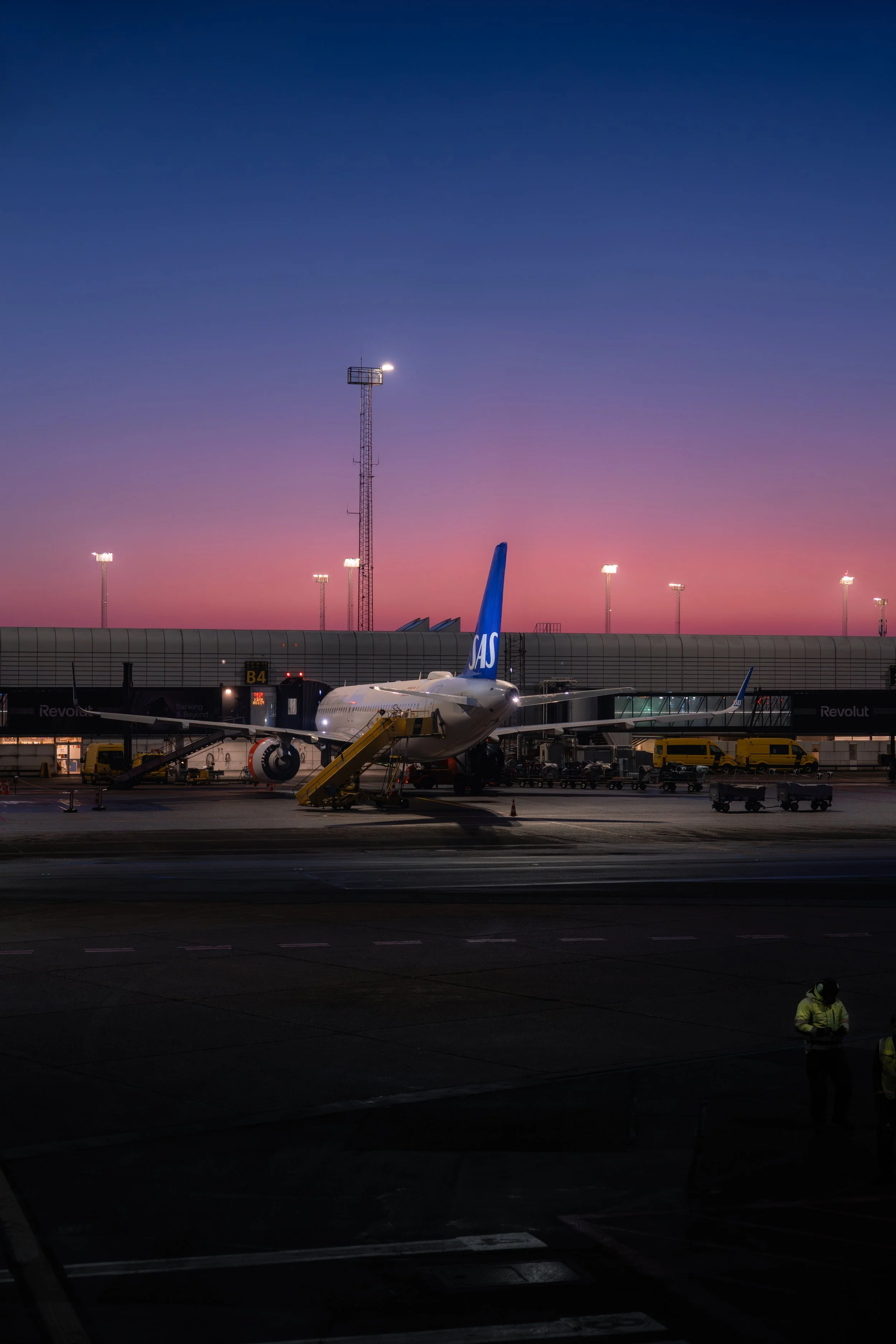An airplane parked at an airport gate during sunset, with a pink and purple sky, airport personnel, and ground service vehicles nearby.