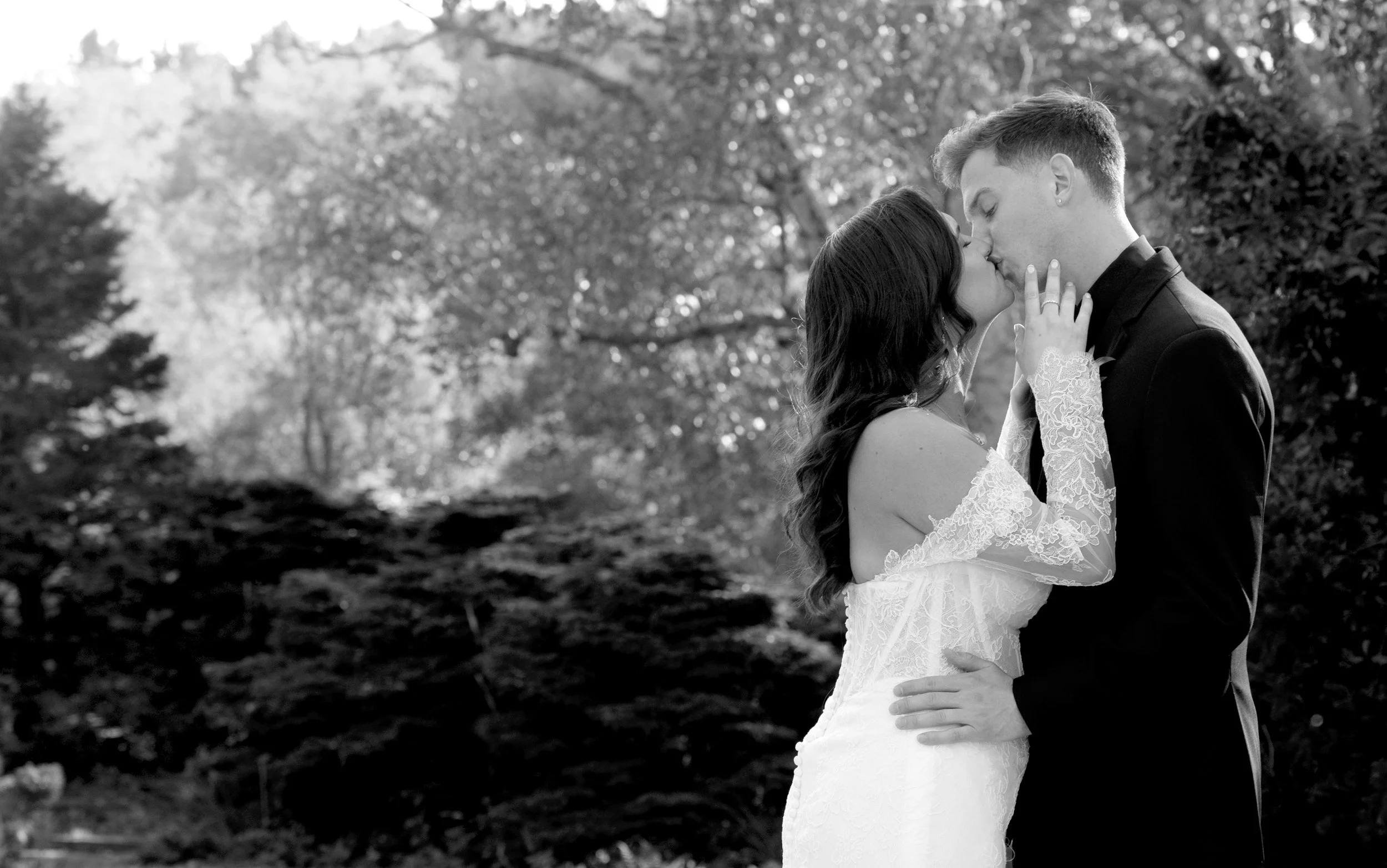 A black and white photo of a couple kissing outdoors. The woman is wearing a white lace wedding dress and the man is in a black suit. They are holding each other close with trees in the background.