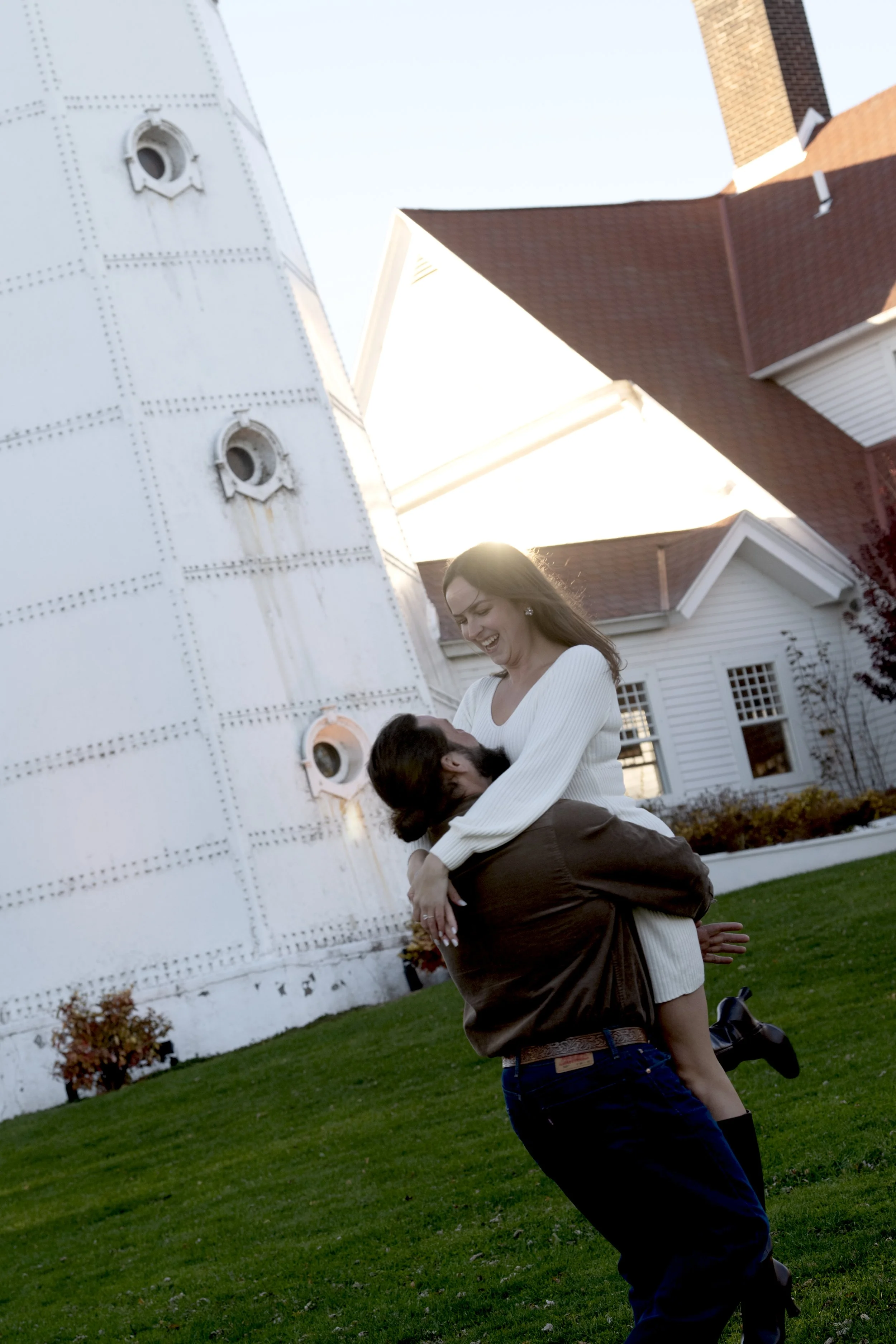 A woman and a man are outdoors at sunset, engaging in a playful moment. The woman is sitting on the man's shoulders, smiling and laughing, while the man is holding her securely. They are in front of a white house with white and red trim, and a tall, 