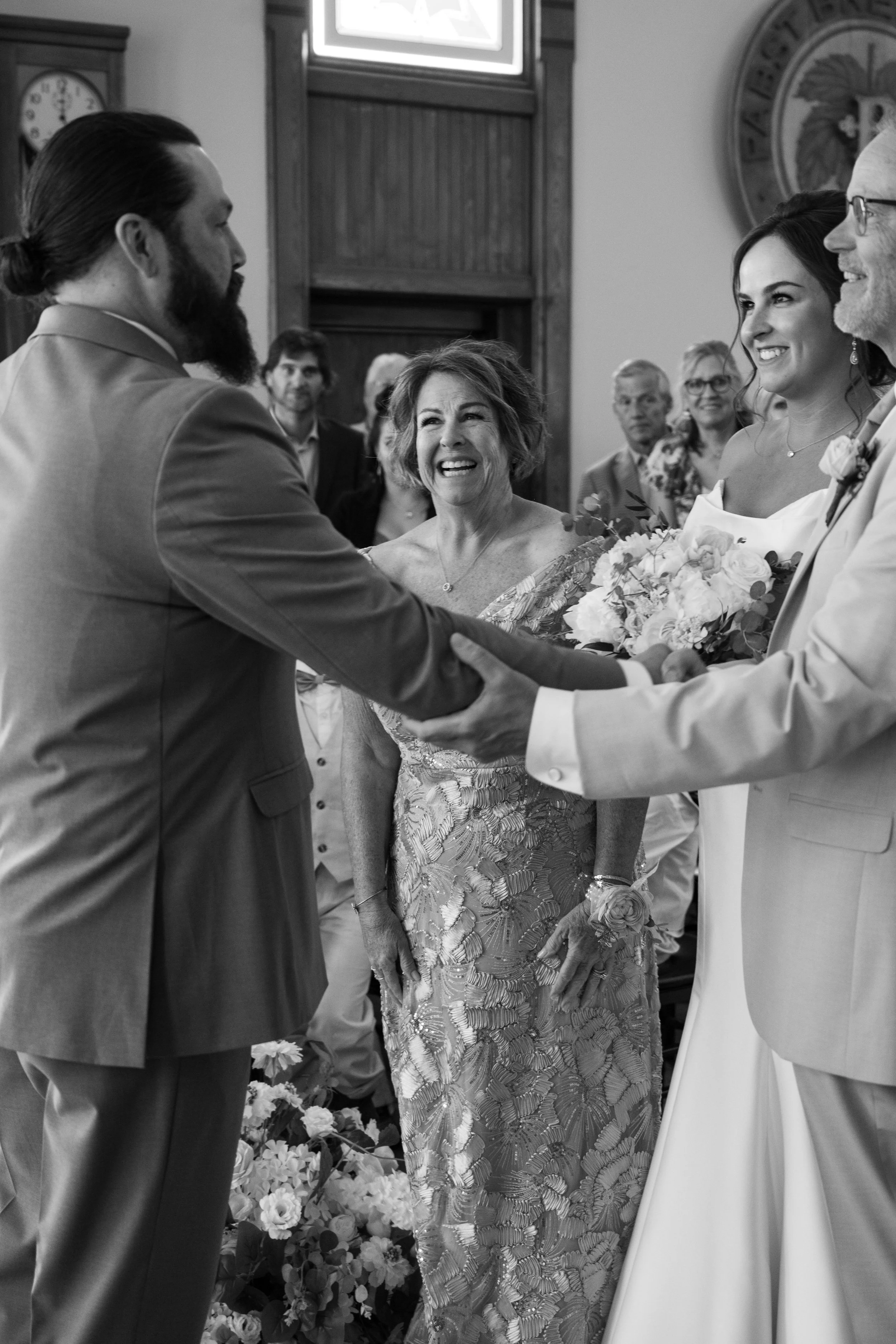 A black and white photo of a wedding ceremony where a woman in a lace dress is smiling at a man in a suit, while another man in glasses holds her hand. In the background, several guests are watching, and the woman is holding a bouquet of flowers.
