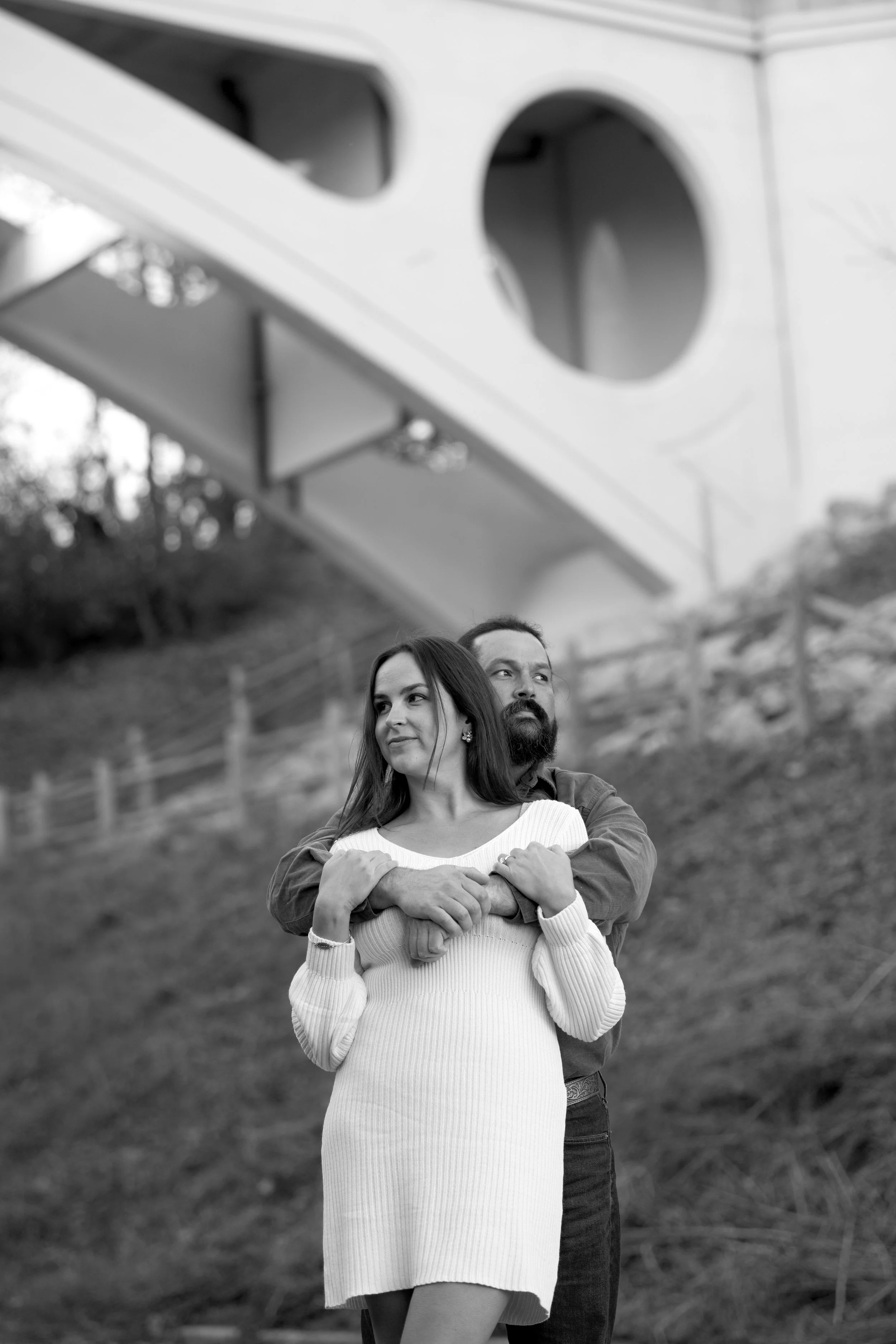 A man and a woman standing outdoors, with the man hugging the woman from behind. The woman is wearing a white sweater dress and the man is wearing a jacket. They are in a scenic area with a grassy hill and a fence in the background.