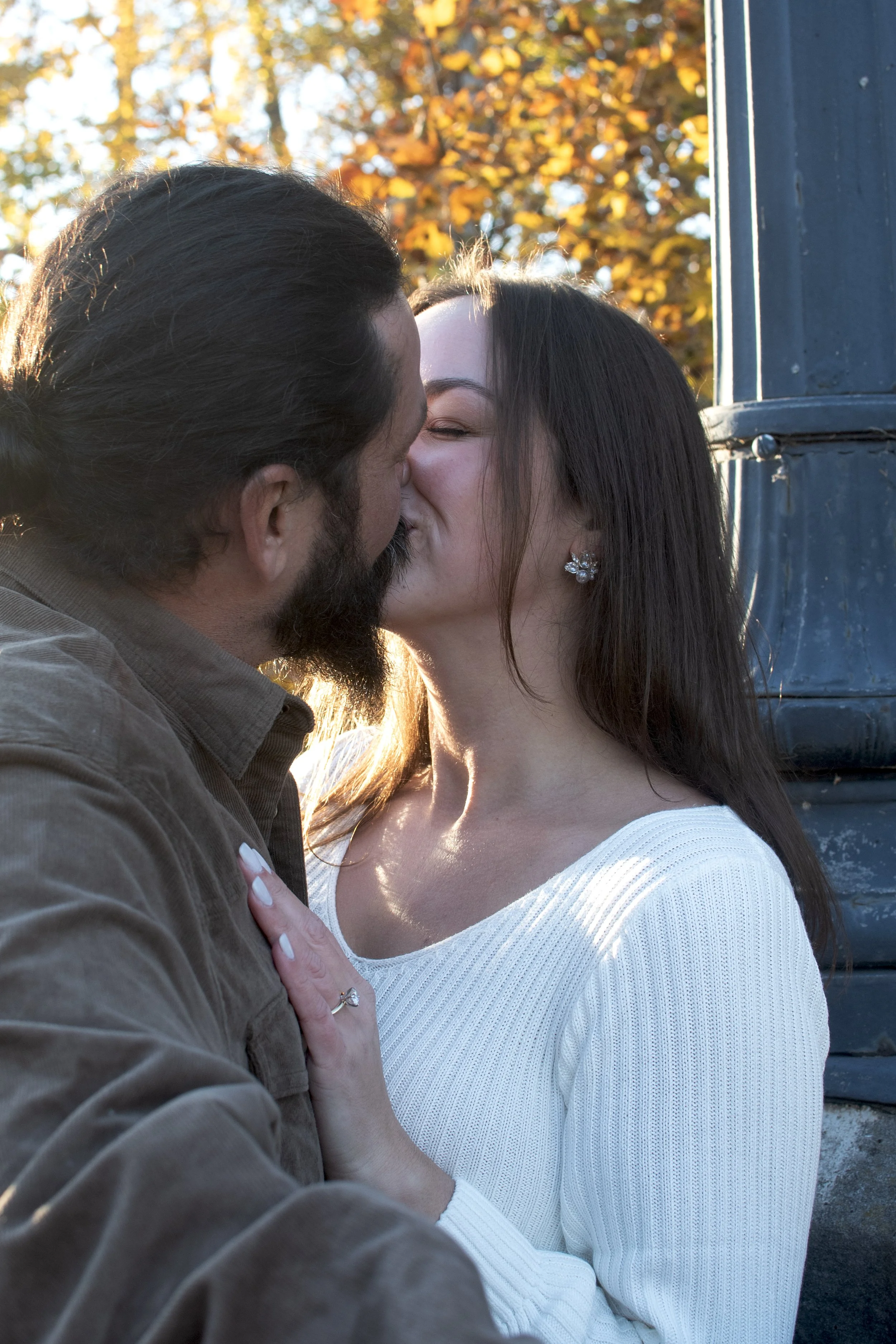 A man and woman are sharing a kiss. The woman has long dark hair, earrings, and is wearing a white sweater. The man has dark hair, a beard, and is wearing a brown jacket. They are outdoors in front of a background of orange autumn leaves and a blue v