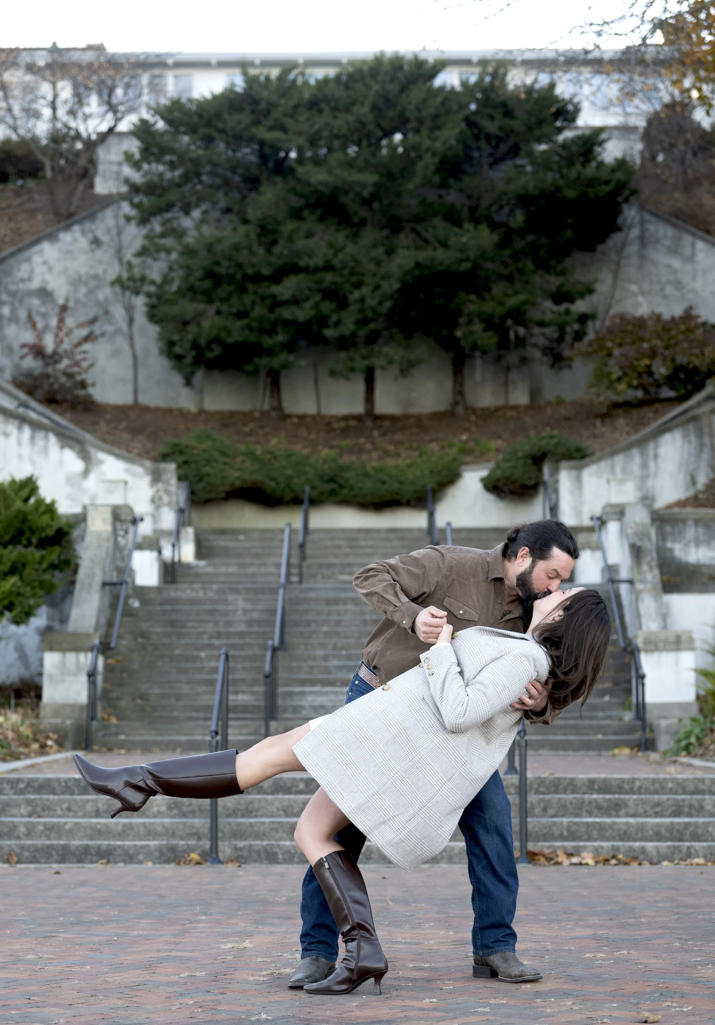 A man and woman dancing on a brick stairway outdoors. The man is dipping the woman, who is leaning back with her legs extended and wearing high boots. The background includes trees, steps, and a white wall.