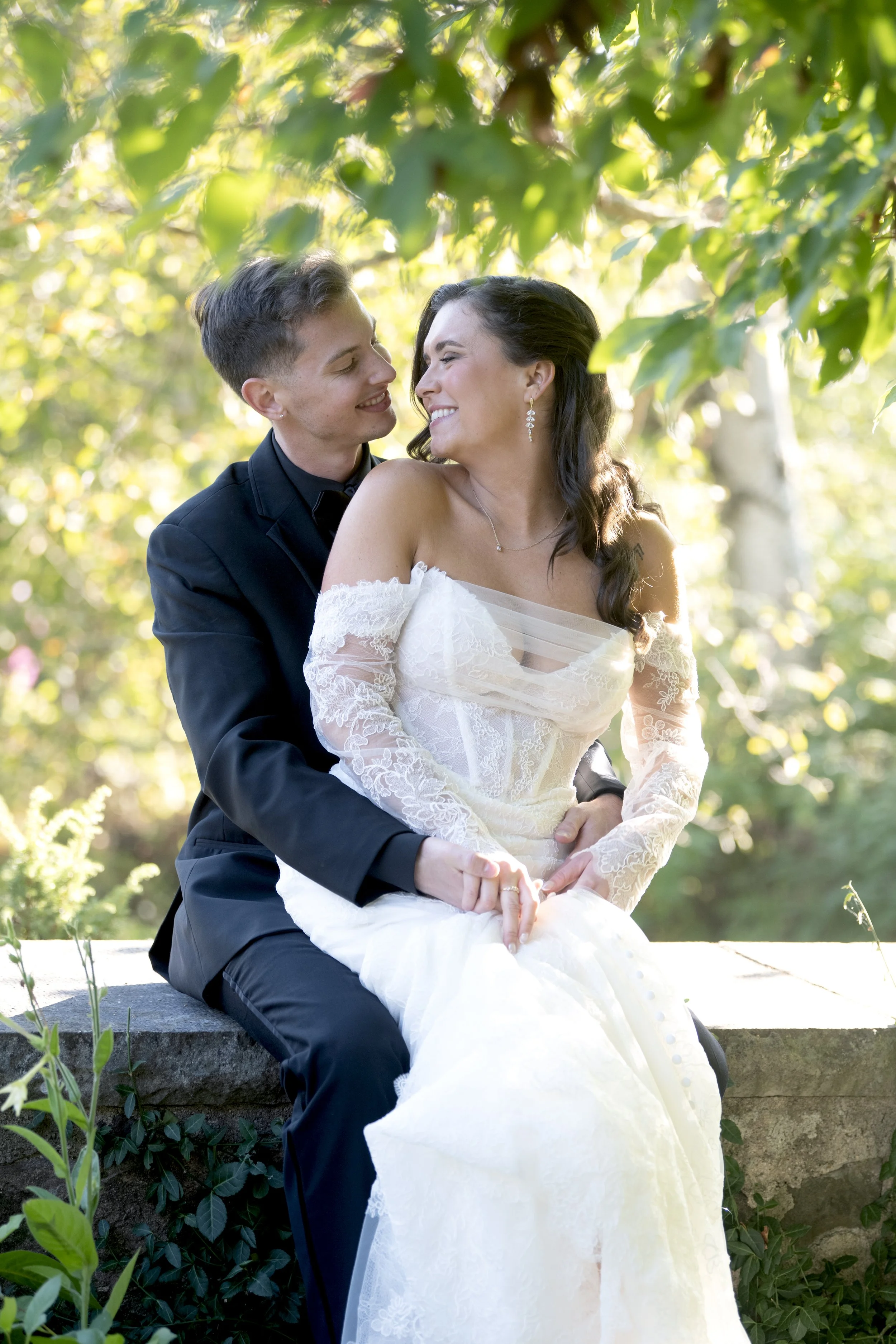 A couple dressed in wedding attire sitting on a stone ledge outdoors, smiling and gazing at each other surrounded by green foliage and dappled sunlight.