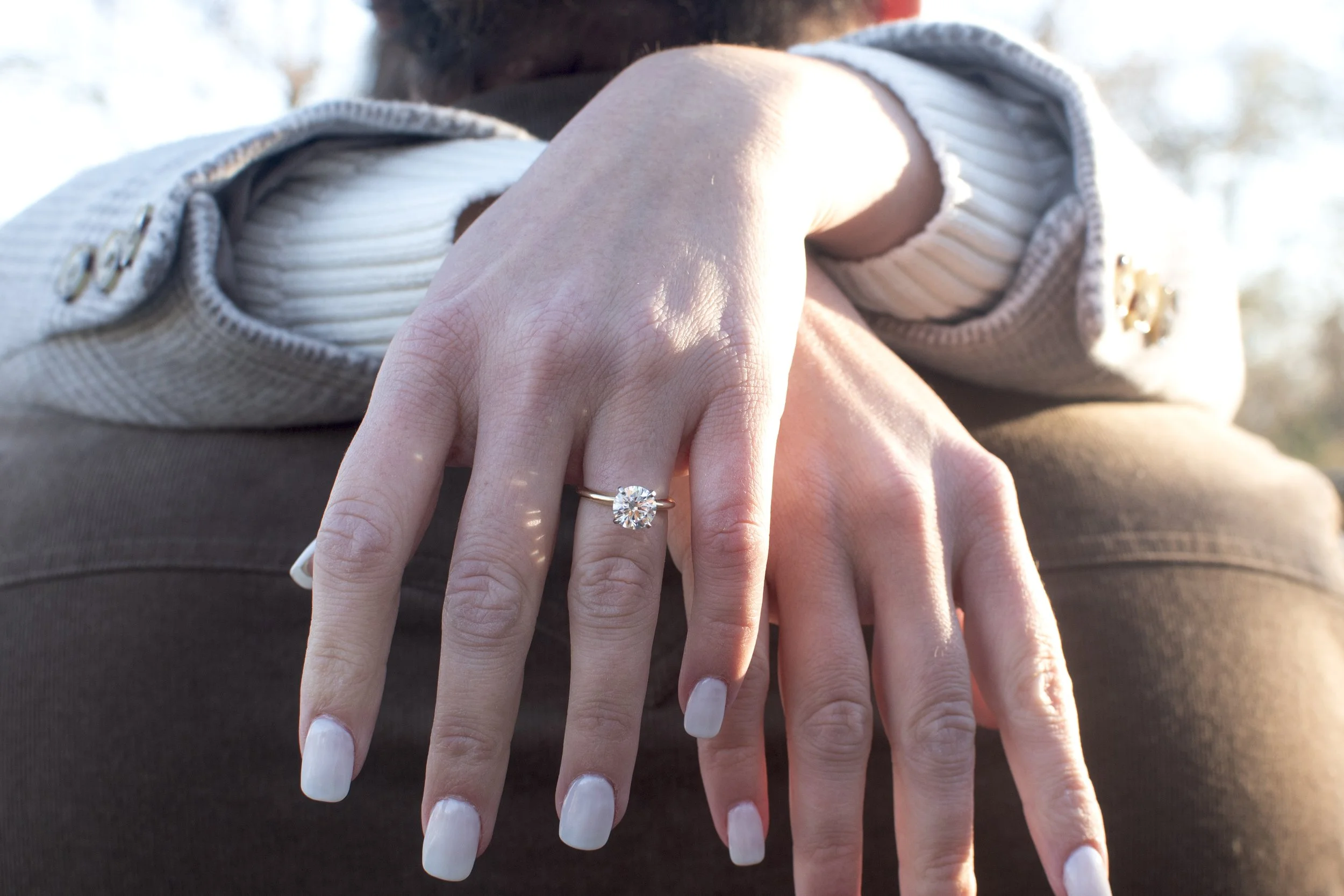 Close-up of two hands, one with a large diamond engagement ring, resting on a person's lap outdoors with sunlight.