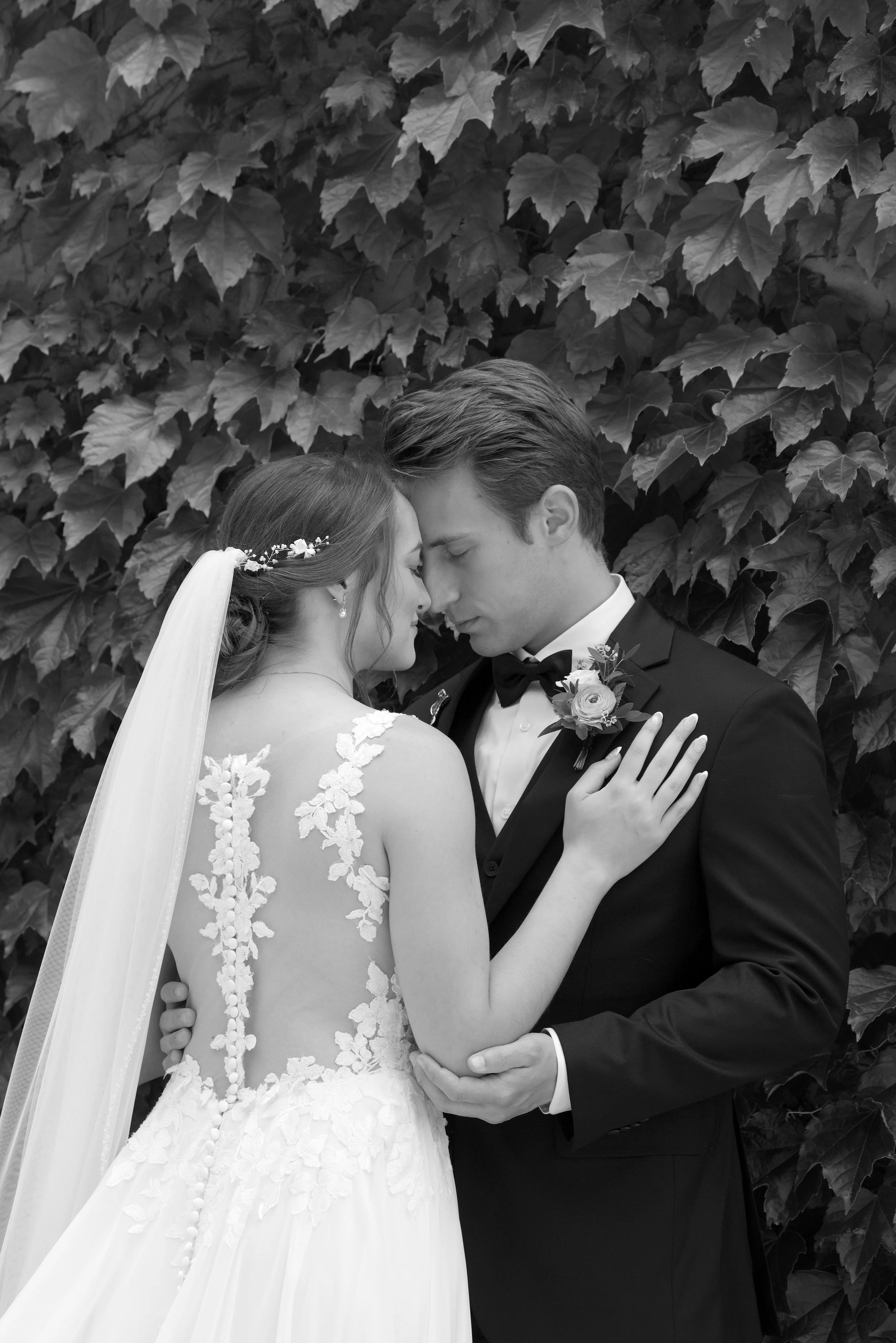 A bride and groom in wedding attire sharing an intimate moment, with their foreheads touching, standing in front of a background of large leafy plants.