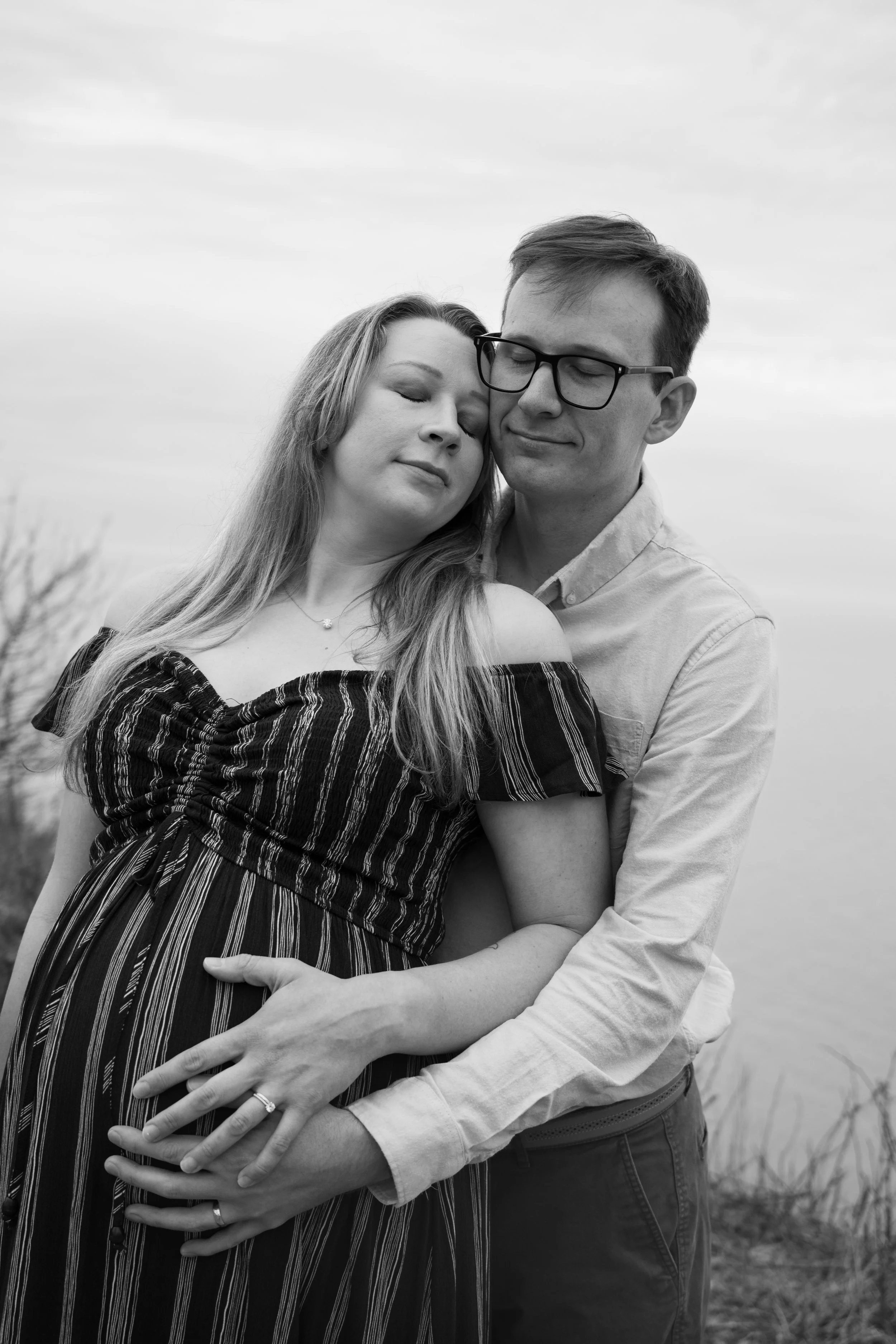 A black and white photo of a couple with their eyes closed, embracing outdoors in a field with cloudy sky in the background.
