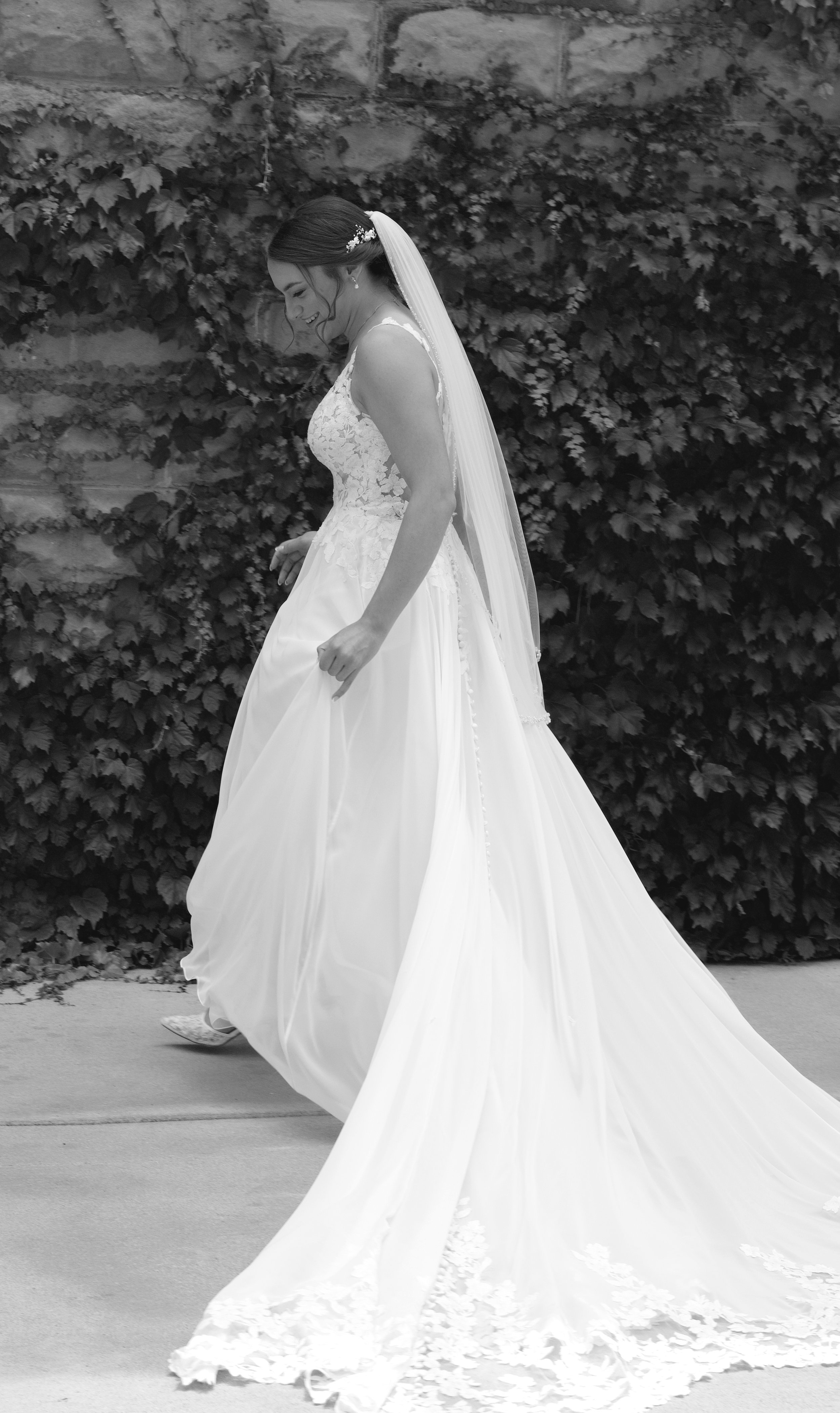 Black and white photo of a smiling bride in a wedding dress with a long train, standing outdoors against a backdrop of leafy vines and stone wall.