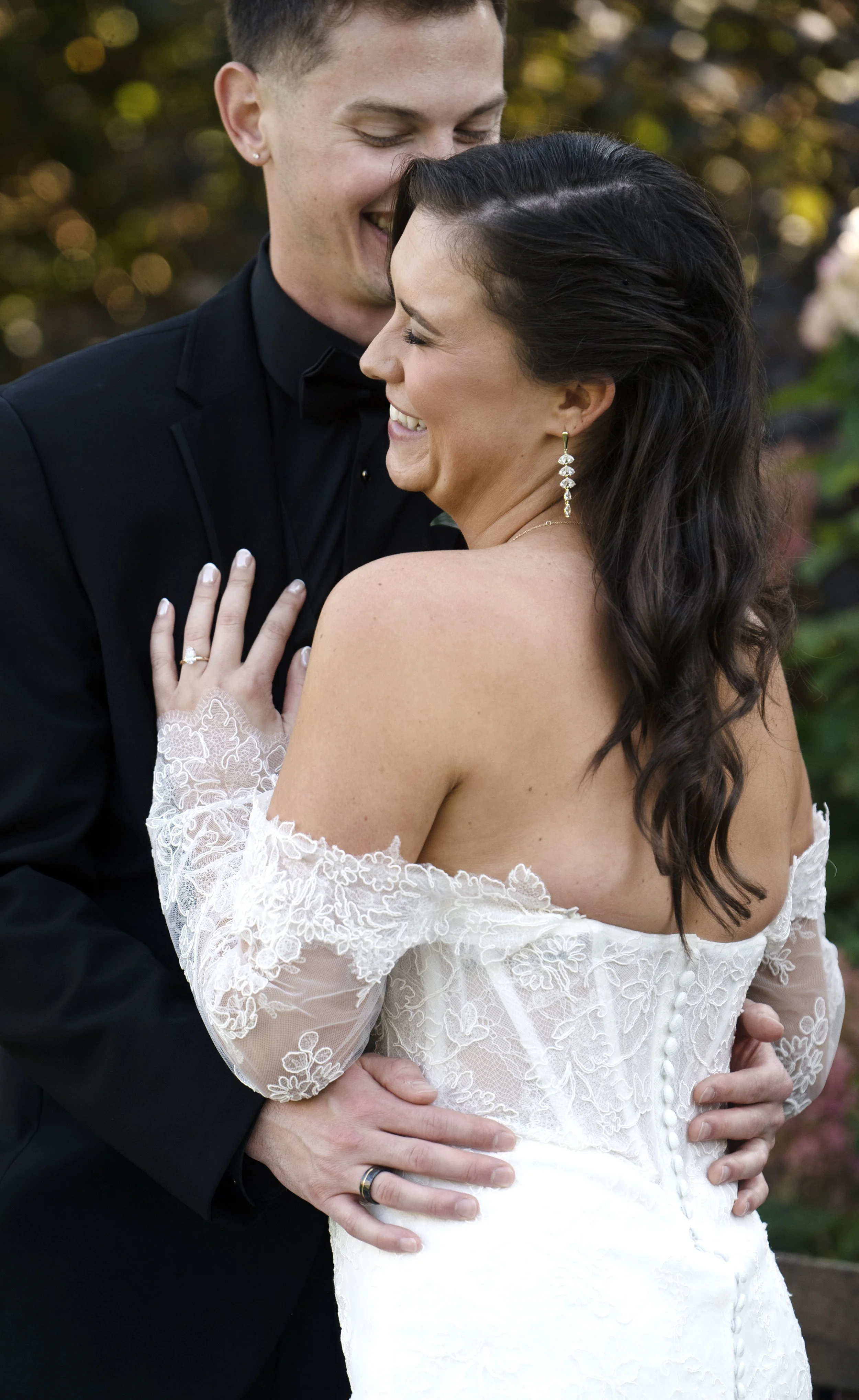 A newlywed couple smiling and embracing outdoors, with the groom dressed in a black suit and the bride in a white lace wedding dress, both showing happiness.