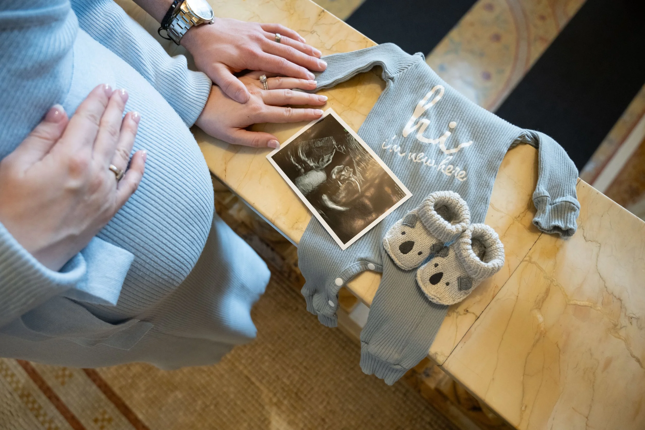 A pregnant person with hands on her belly, wearing a light blue sweater, sitting at a beige marble table. On the table are tiny knitted baby socks, a gray onesie with white writing, and an ultrasound picture. The setting appears to be a cozy indoor space.