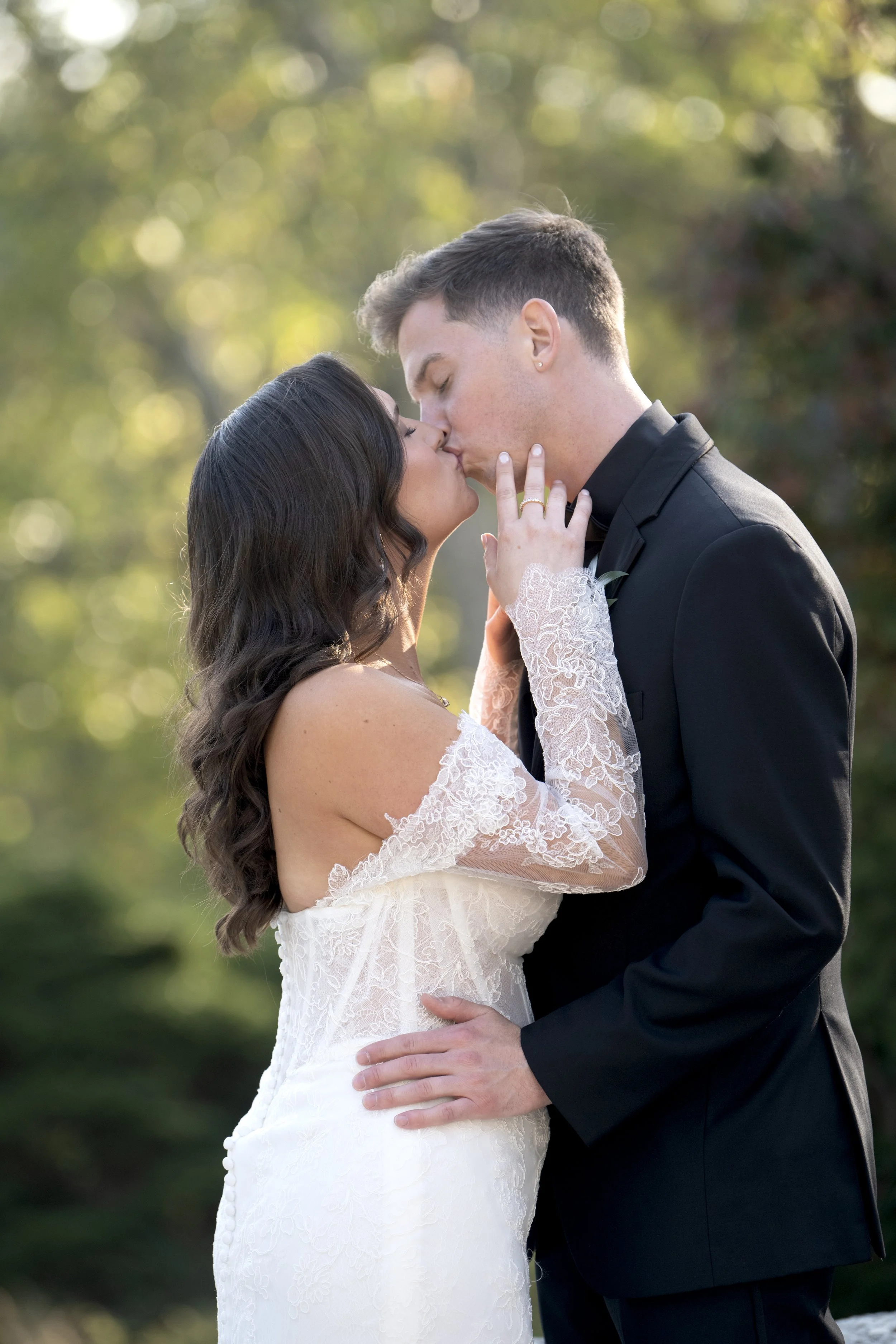 A bride and groom kissing outdoors, with the bride wearing a white lace dress with long sleeves and the groom in a black suit, surrounded by a blurred background of green trees and soft sunlight.