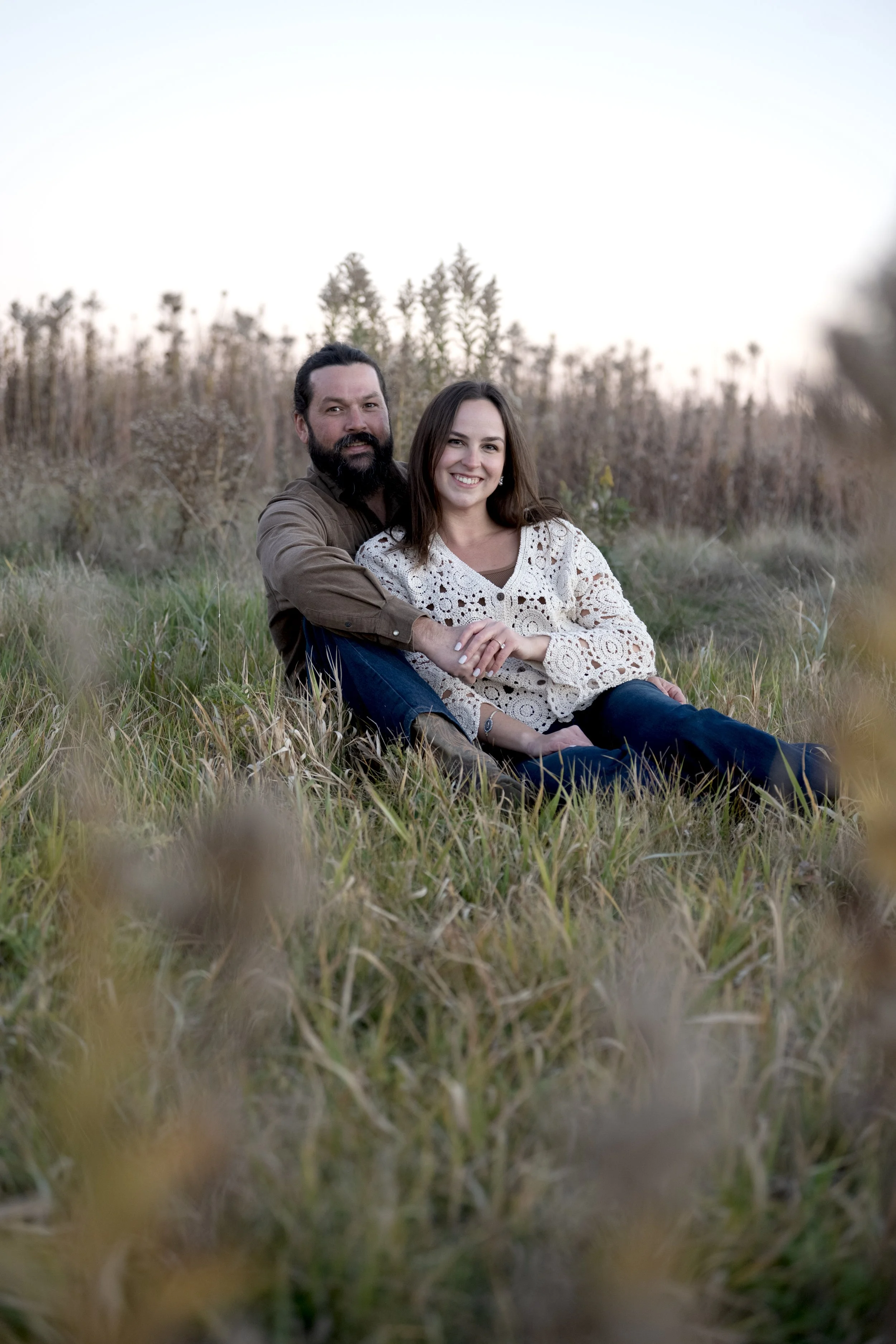 A man with dark hair and a beard and a woman with long brown hair sitting in a grassy field during sunset, smiling at the camera.