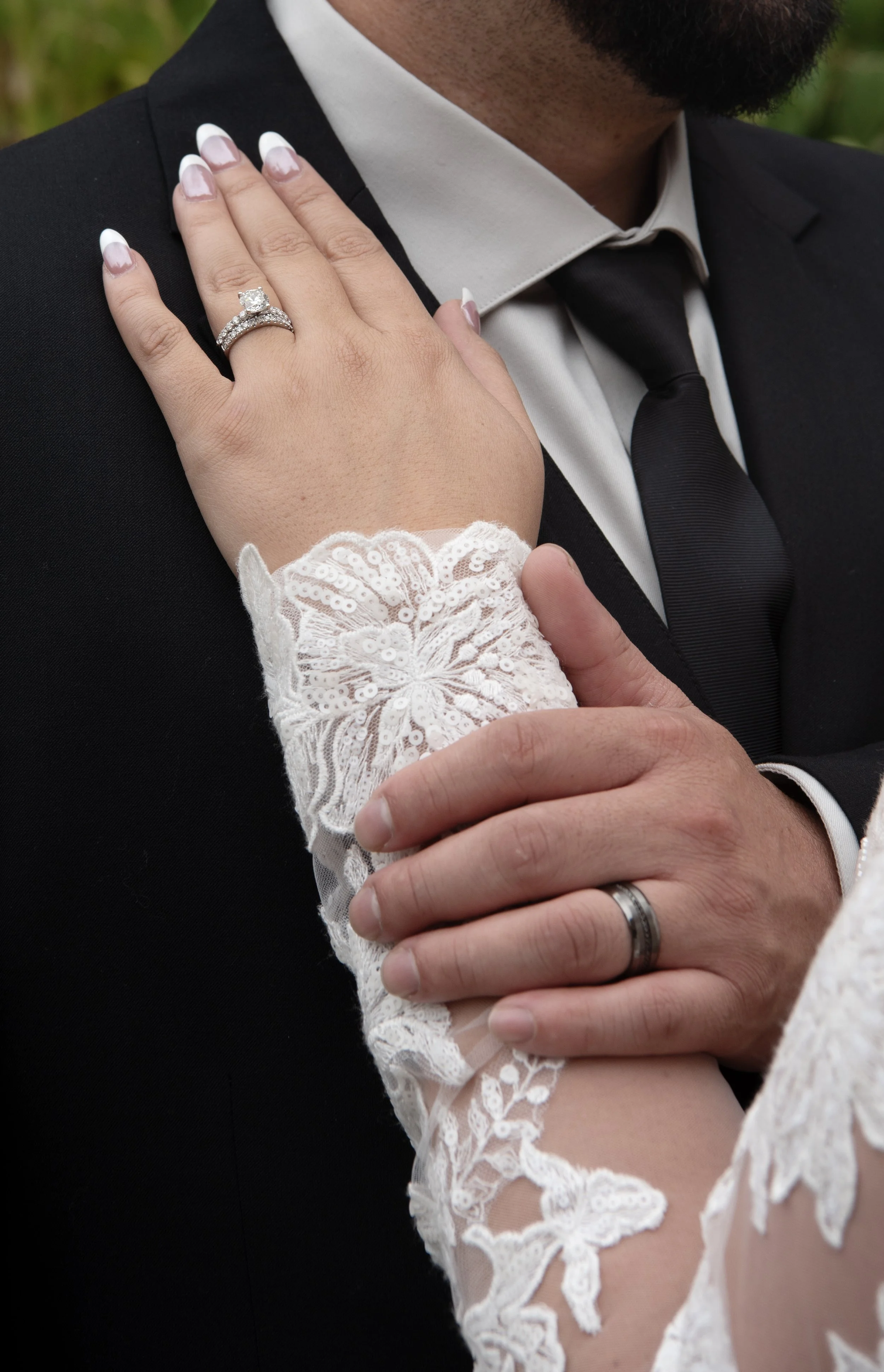 Close-up of a woman's hand with an engagement ring and wedding band on her finger, resting on a man's shoulder, who is dressed in a black tuxedo and necktie. The woman is wearing a white lace dress with long sleeves.