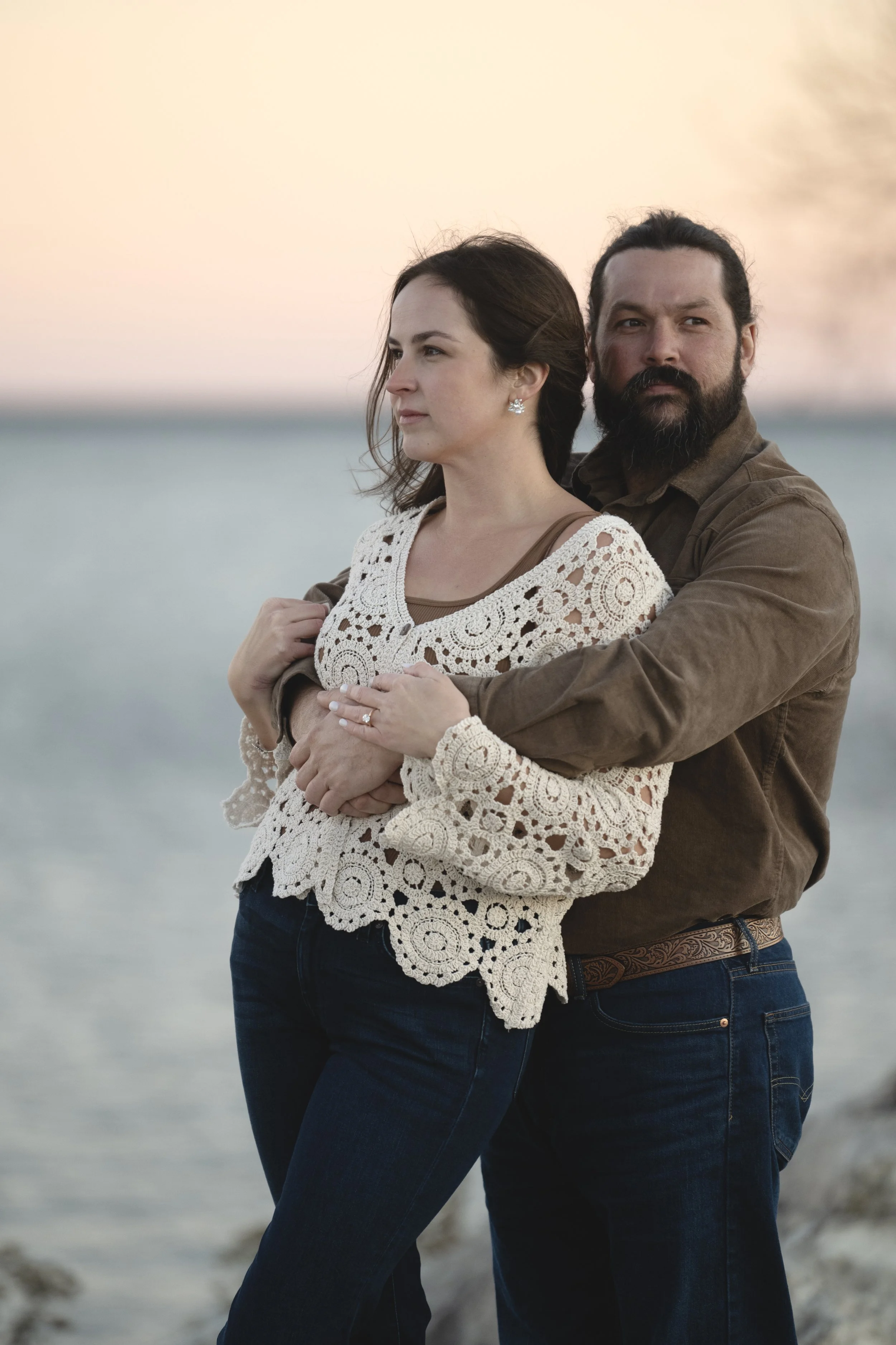 A man and woman standing outdoors at the beach, facing opposite directions, with the ocean behind them at sunset.