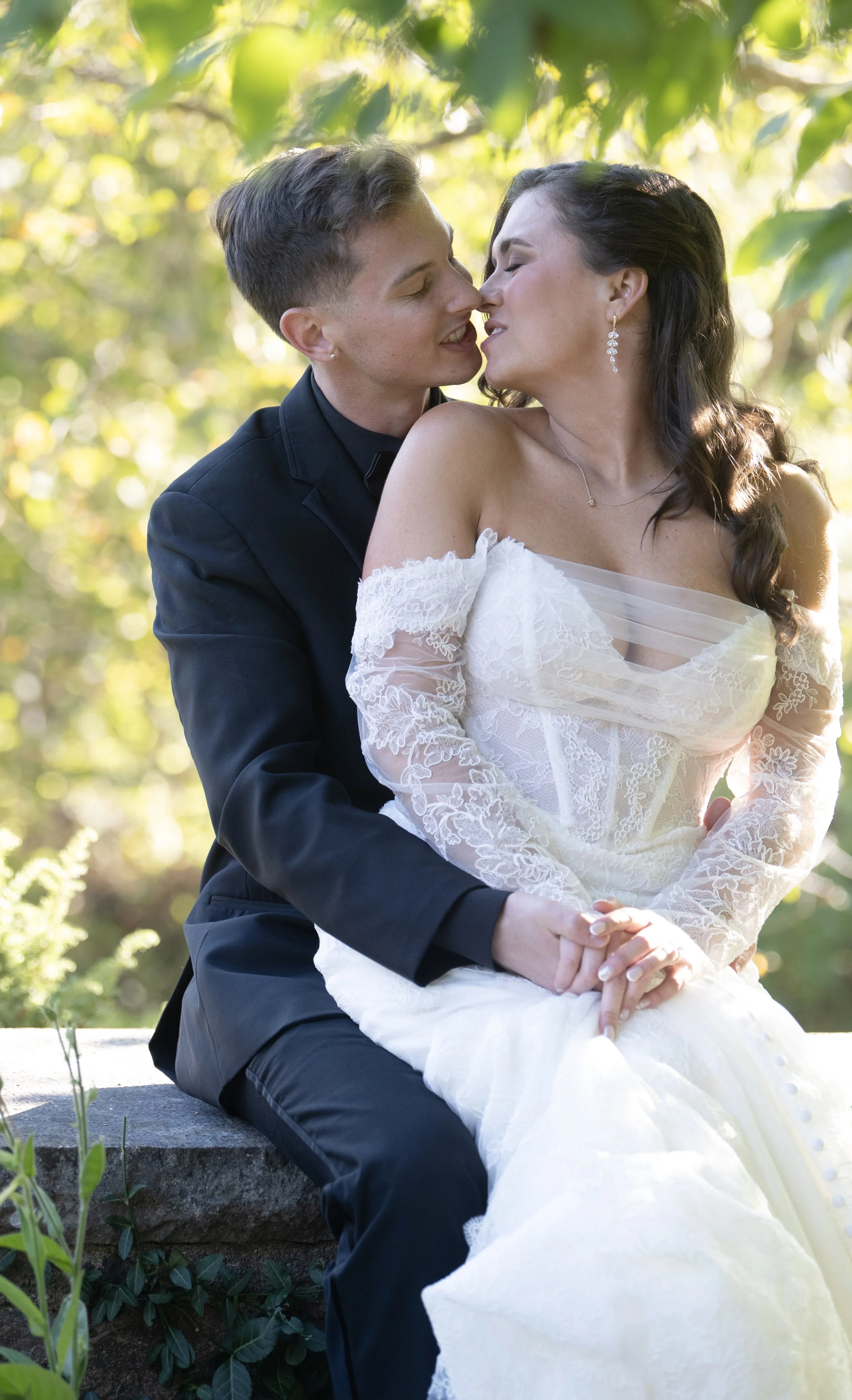 A couple, dressed in wedding attire, sitting closely outdoors in a lush, green setting, with their faces nearly touching and eyes closed, sharing an intimate moment.