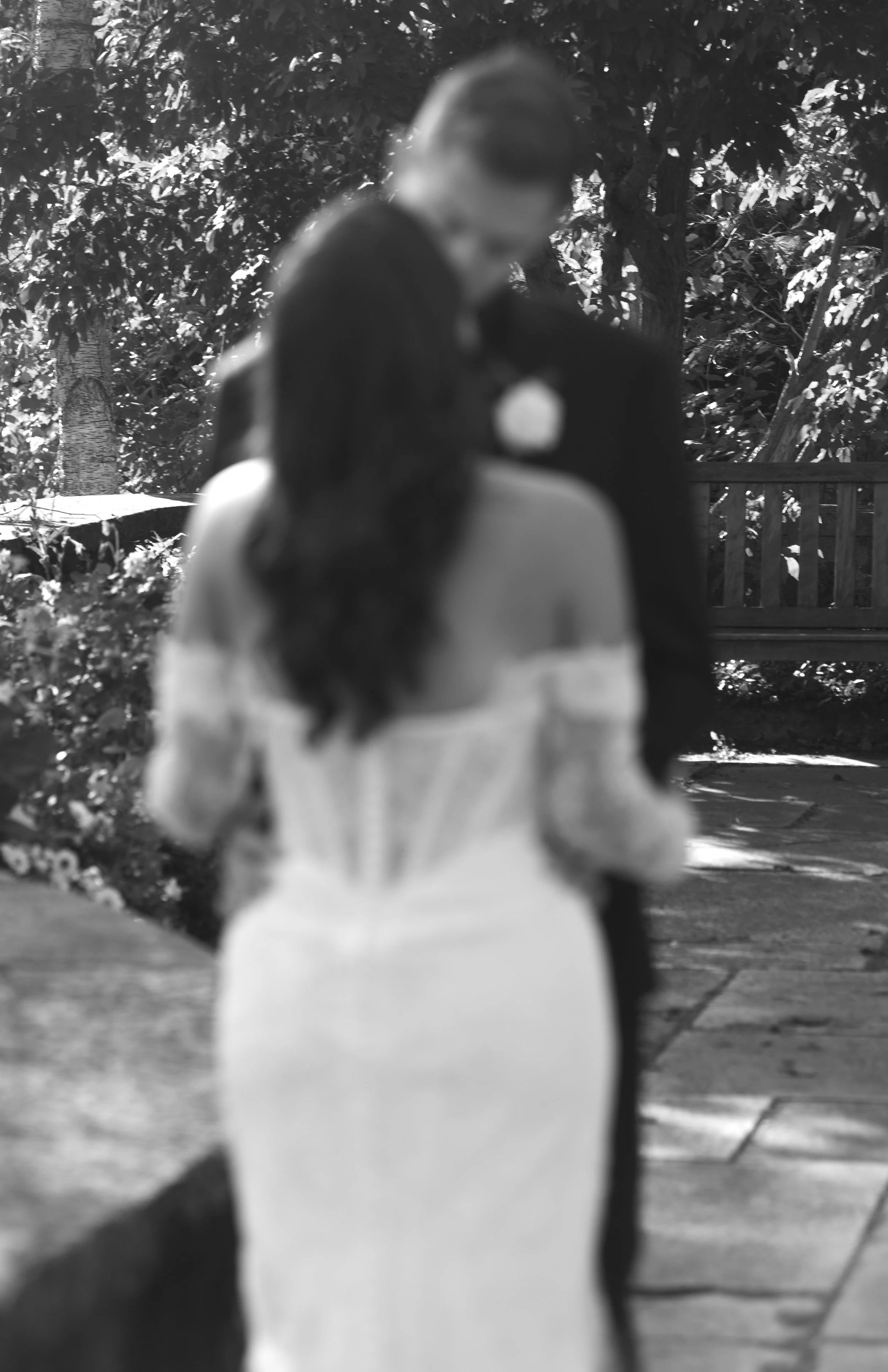 A black and white photograph of two women embracing outdoors, with trees and a wooden fence in the background. One woman in a dark outfit and the other in a light-colored dress.