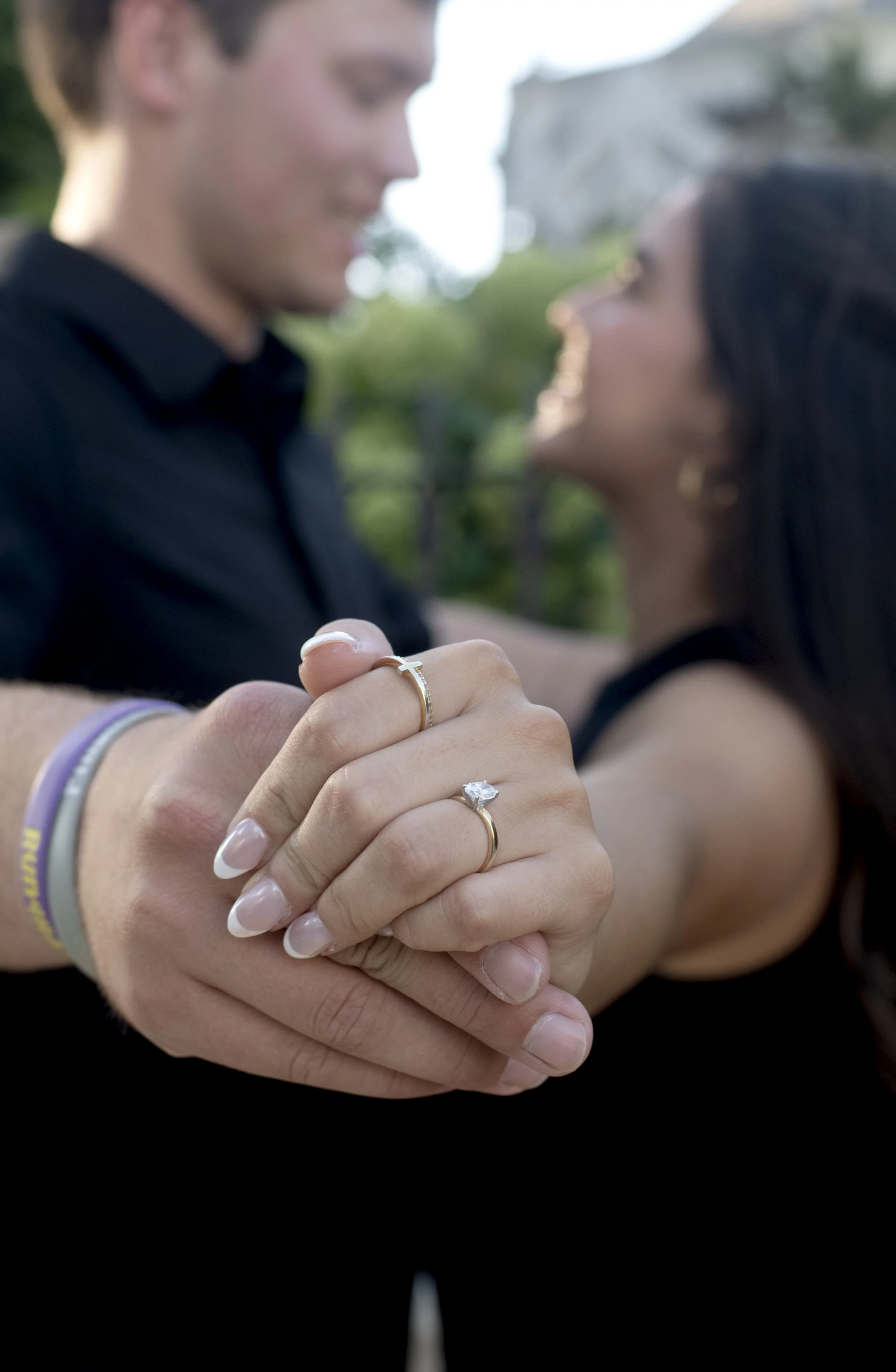 Close-up of hands holding each other, showing wedding and engagement rings, with a blurred couple smiling in the background.