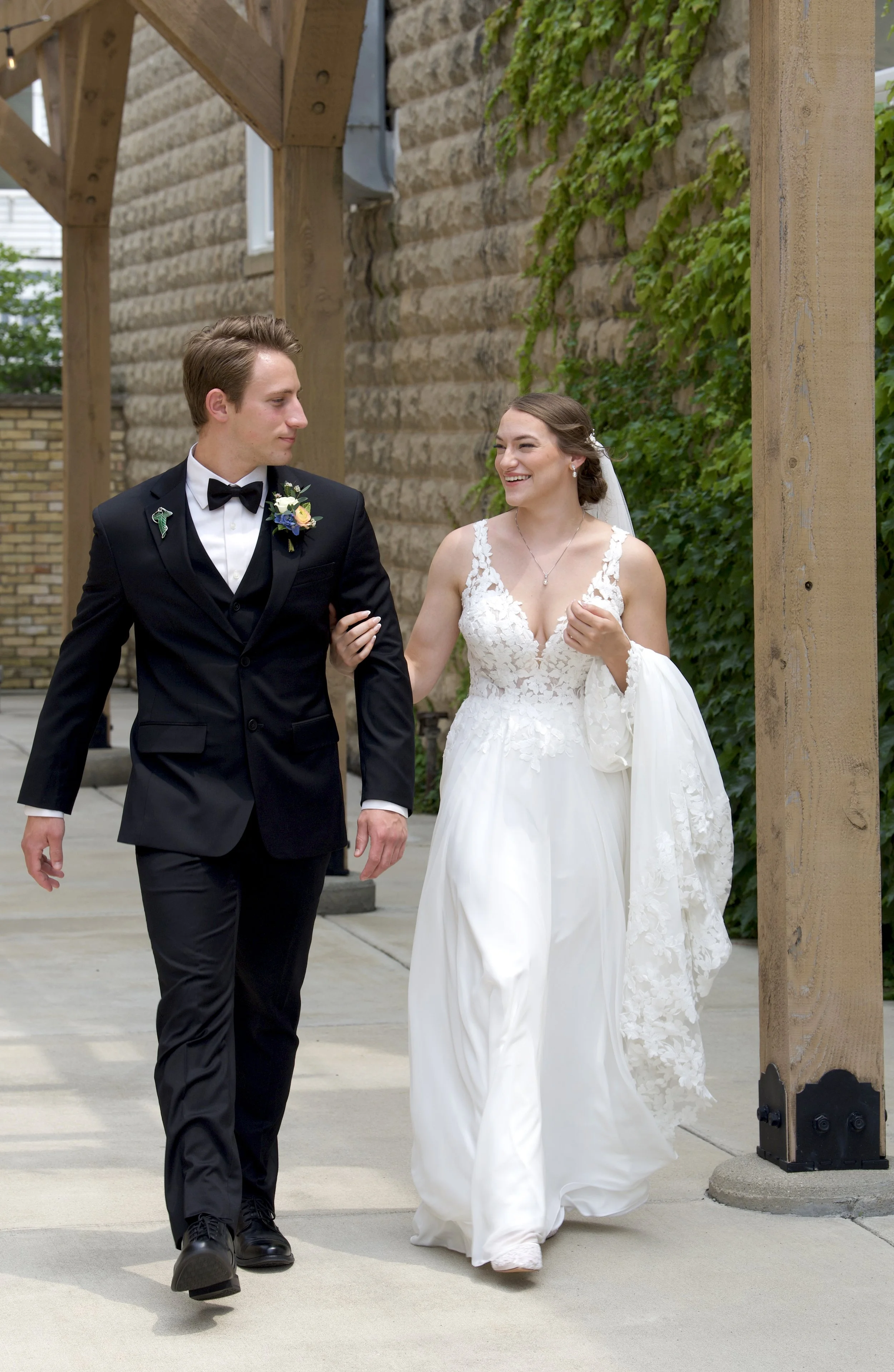 A bride and groom walking arm in arm outdoors, smiling and looking at each other. The groom is dressed in a black tuxedo with a bow tie and boutonniere, and the bride is wearing a white wedding gown with lace details and a veil. They are under a wood