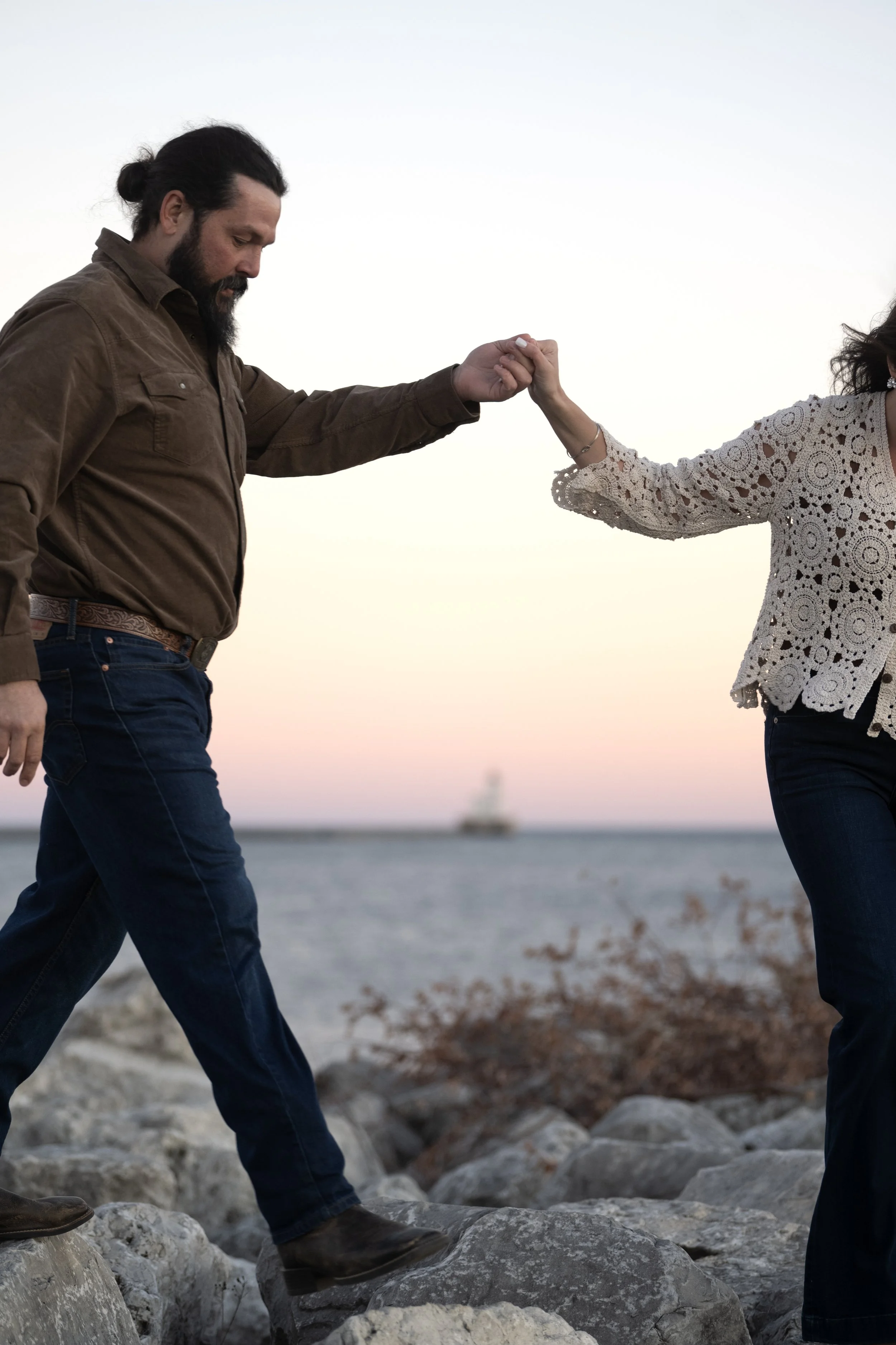 A man and woman holding hands while walking on rocks near a body of water at sunset.