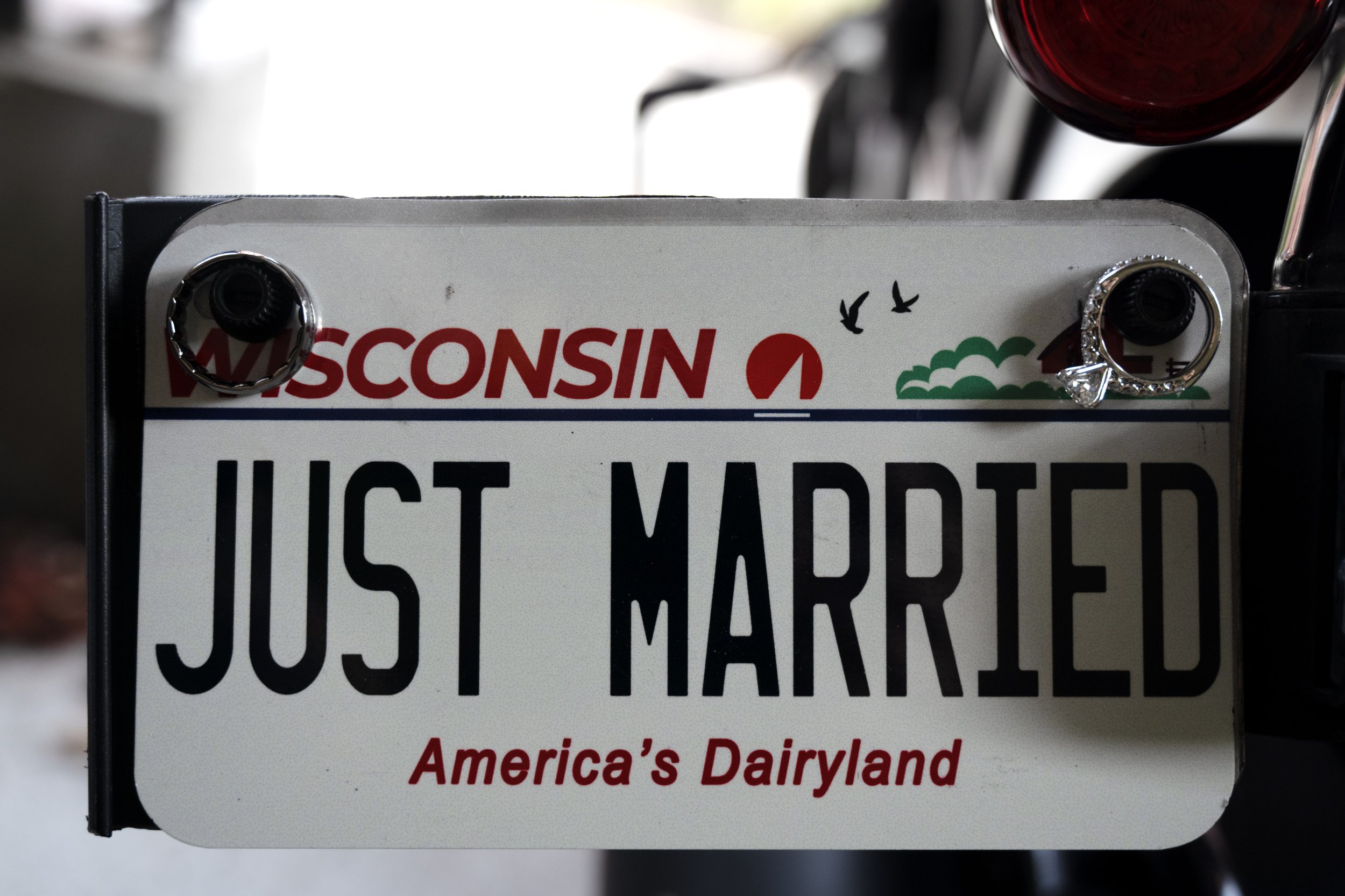 Close-up of a US license plate with the word 'JUST MARRIED' on it, mounted on a vehicle. The plate is surrounded by a decorative ring with rhinestones. The top of the plate features the word 'WISCONSIN' in red letters, and an illustration of birds, t
