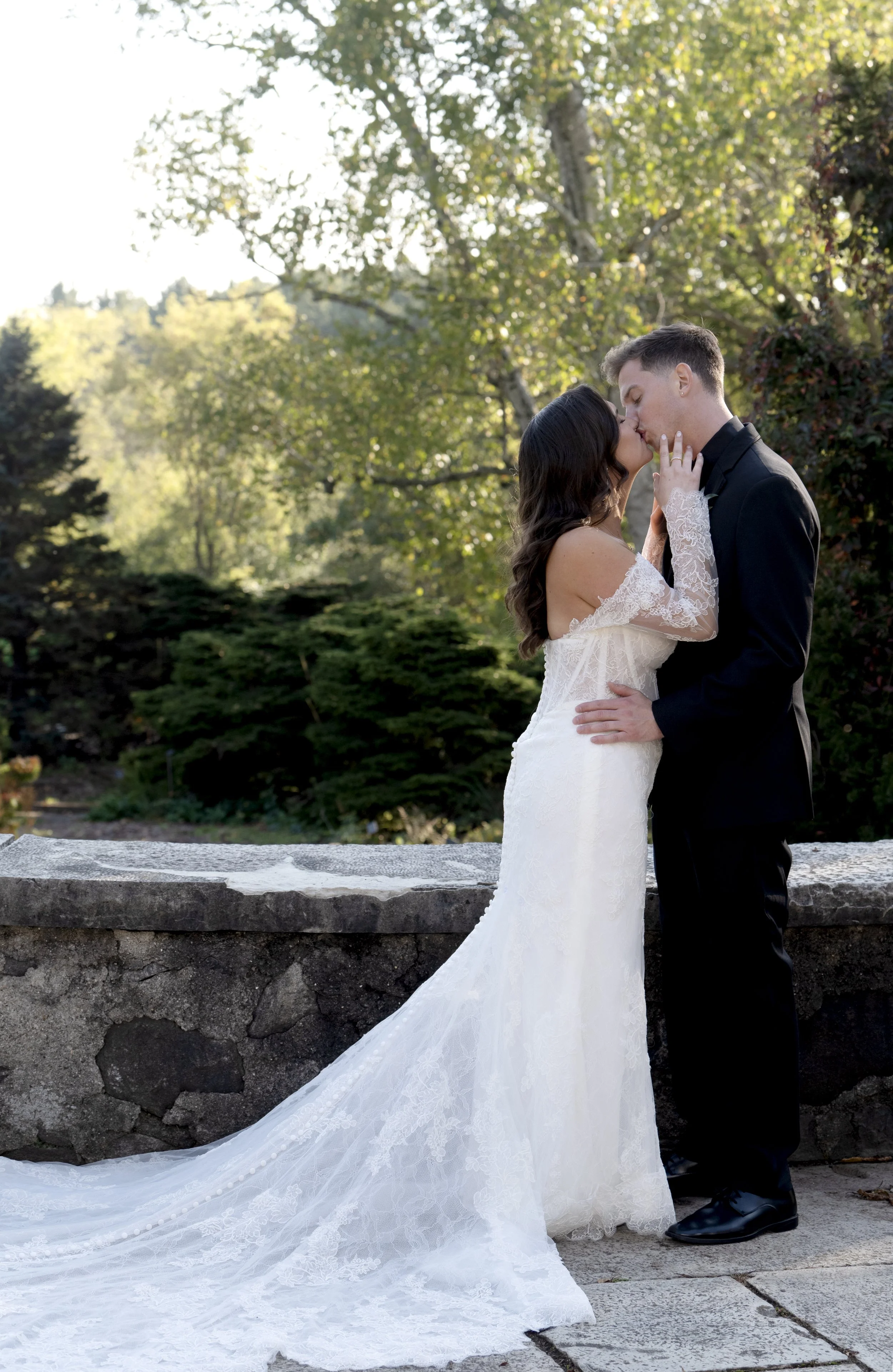 A bride and groom kissing outdoors on a sunny day, with the bride in a white lace wedding gown and the groom in a black suit, standing on a stone pathway with green trees in the background.