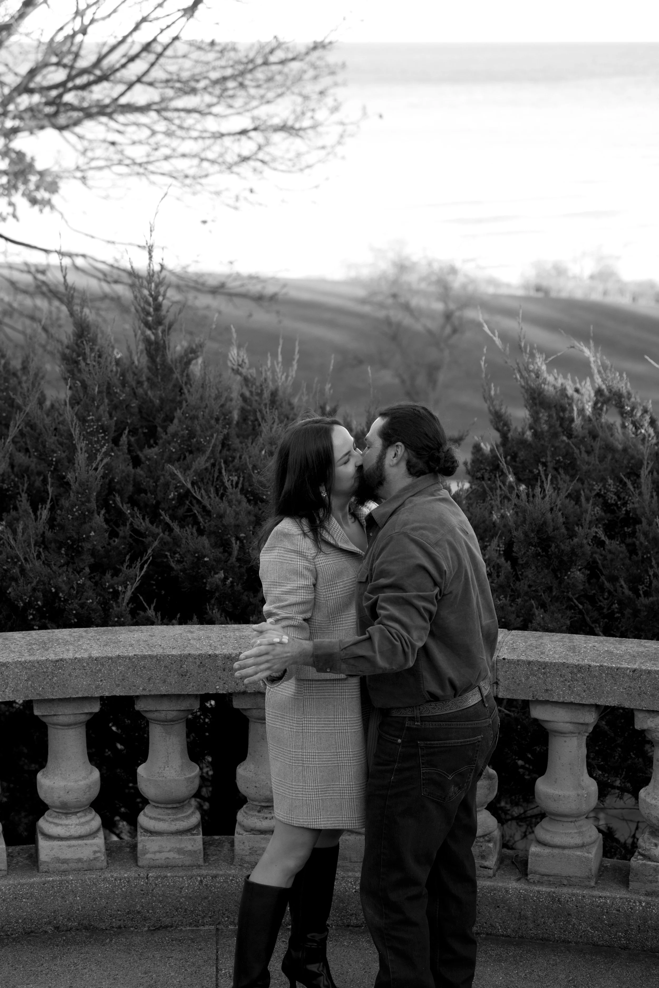 A couple is standing close together and kissing in front of a stone railing with trees and rolling hills in the background.