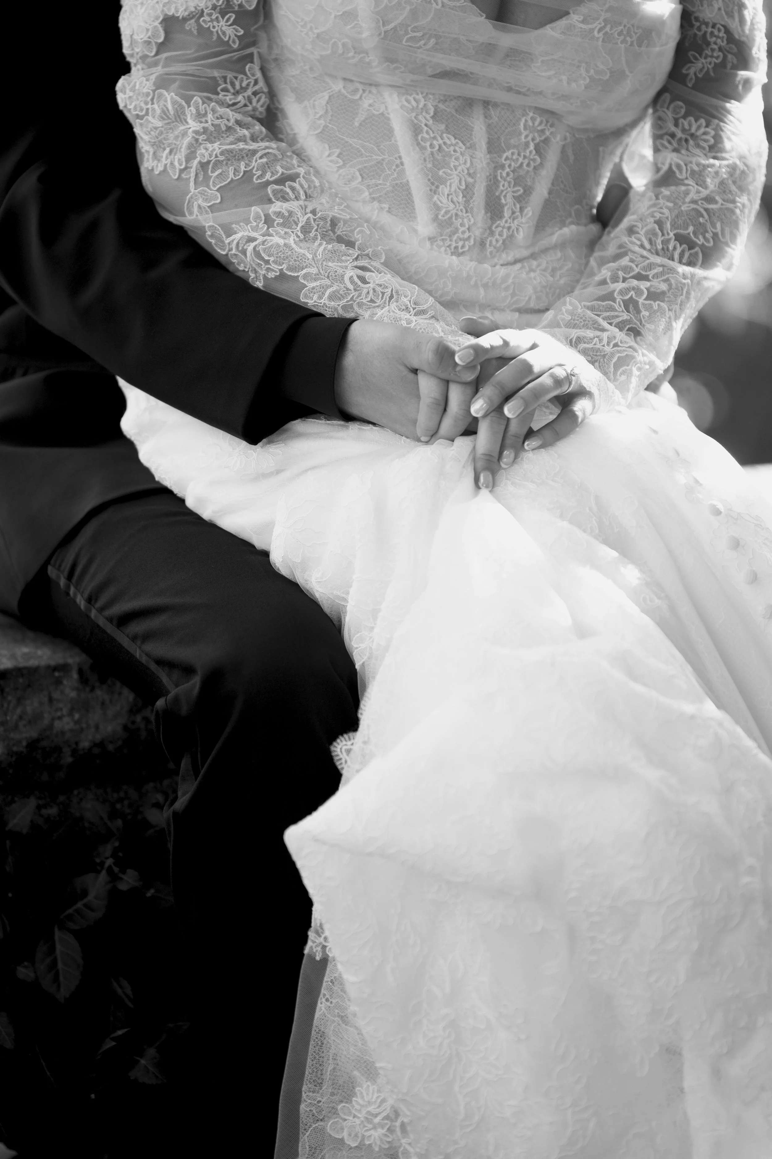 A close-up black and white photograph of a couple's hands gently clasped, with the woman's hand resting atop the man's. The woman is wearing a lace wedding dress with intricate floral embroidery, and the man is dressed in a dark suit. The focus is on