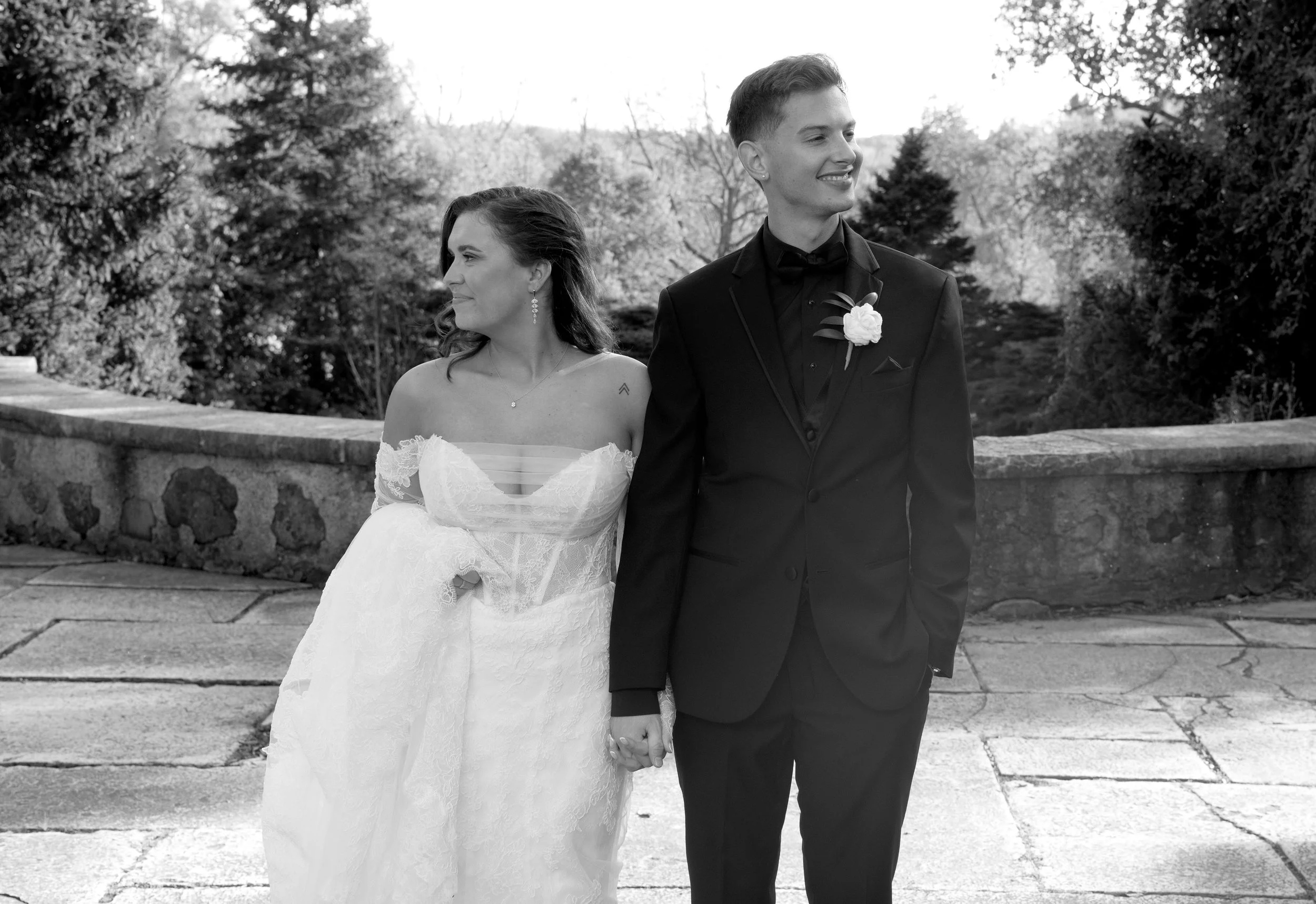 Black and white photograph of a bride and groom holding hands, walking outdoors with trees and a stone wall in the background. The bride is wearing a lace wedding dress and the groom is in a black tuxedo with a boutonnière. They are smiling and looki