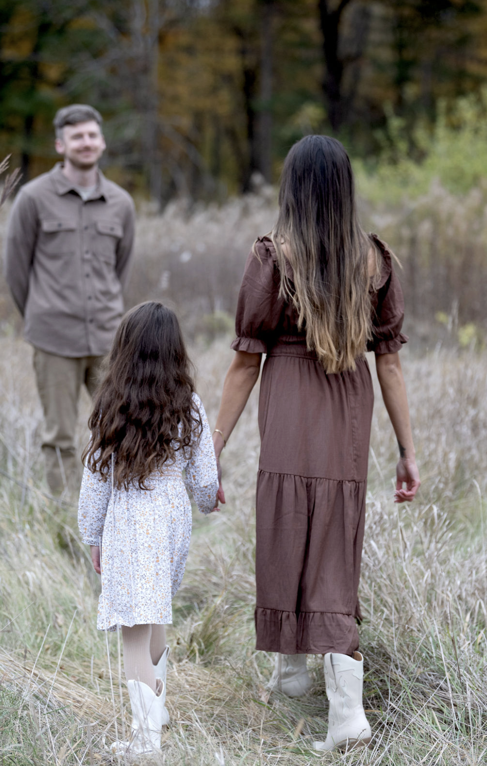 A woman holding hands with a young girl walking through a grassy field with a man standing in the background, surrounded by trees in an outdoor setting.