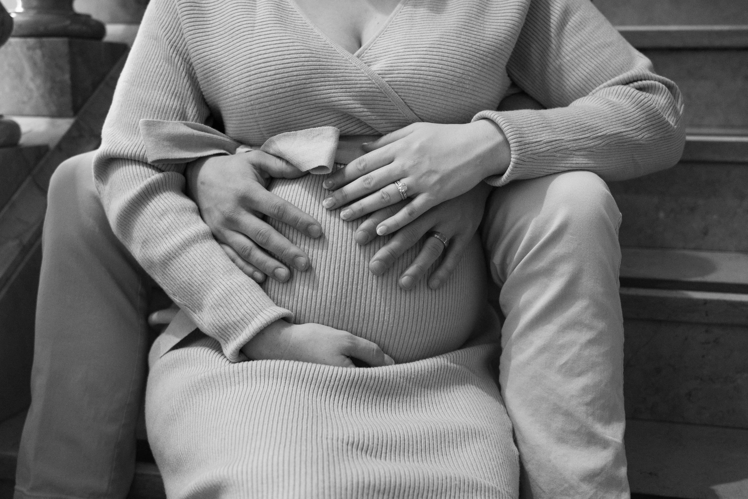 Close-up black and white photo of two people with their hands forming a heart shape on an individual's stomach, sitting on a bench.
