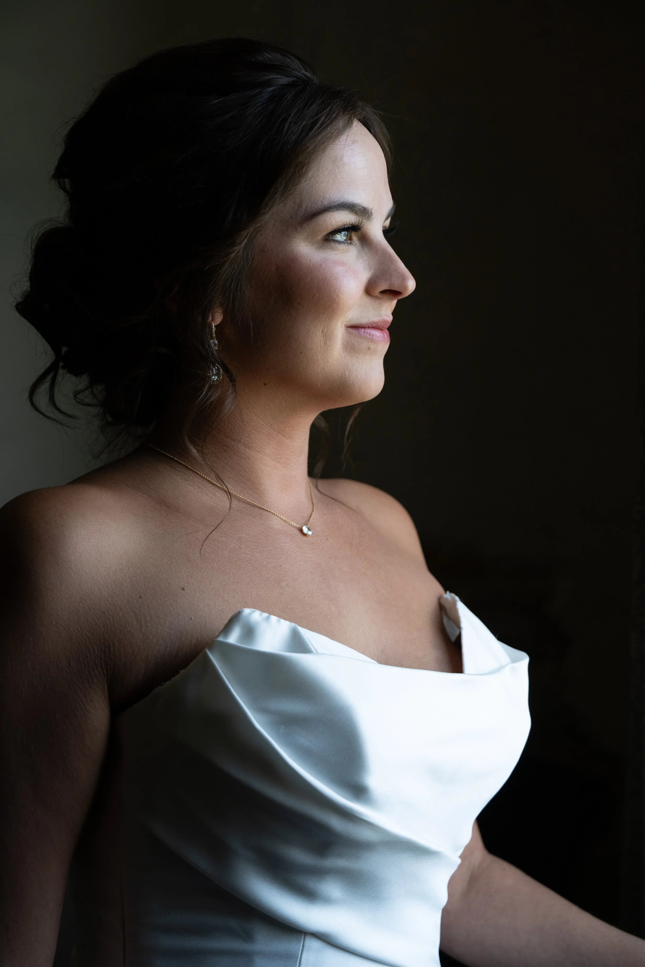 Profile of a woman with dark hair in an elegant updo, wearing a white satin dress with off-the-shoulder ruffles, gold jewelry, and a slight smile, against a dark background.