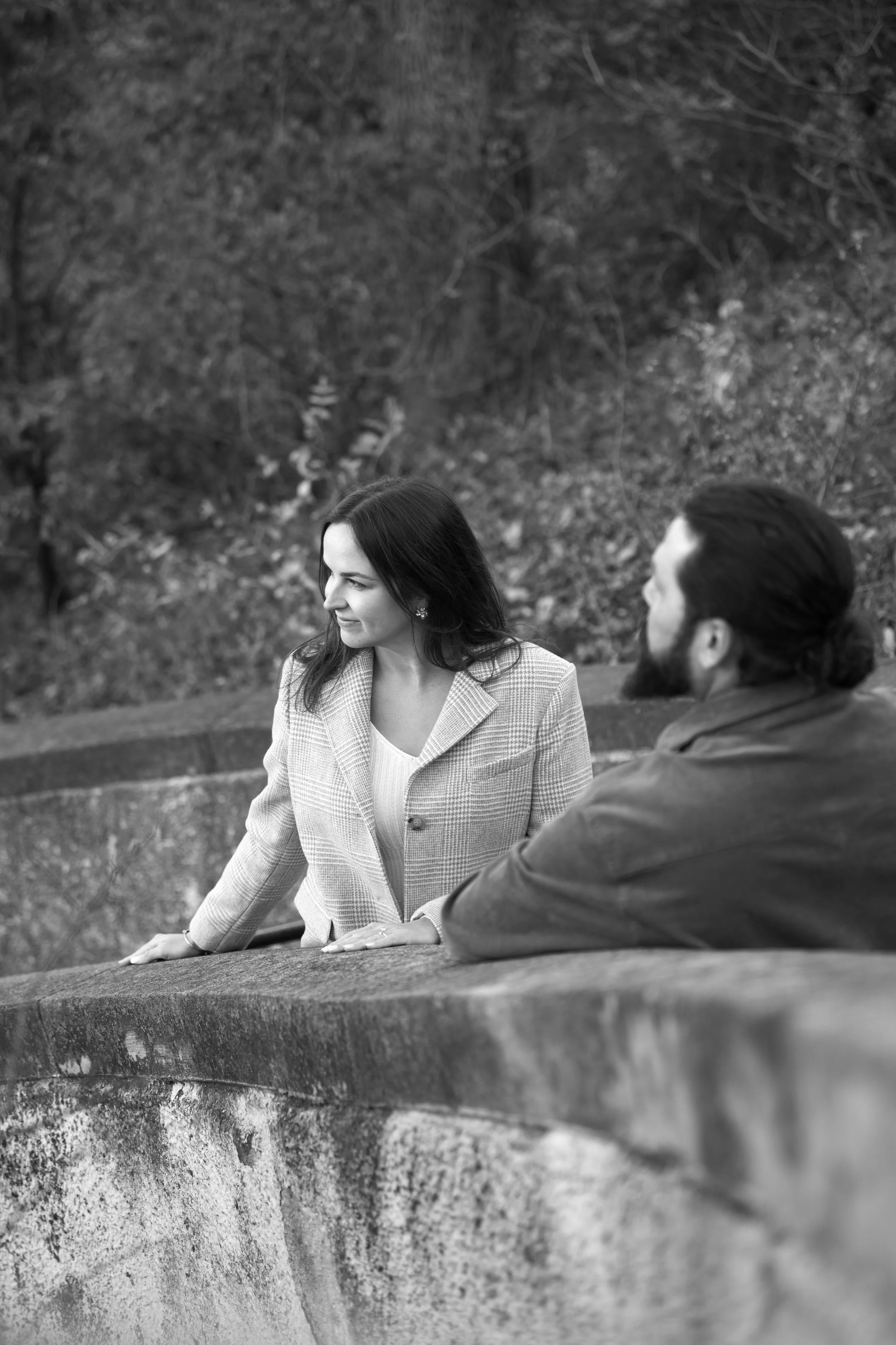 A black and white photo of a woman with long dark hair and a man with a beard and short hair, sitting outdoors on a stone ledge, surrounded by trees and nature.